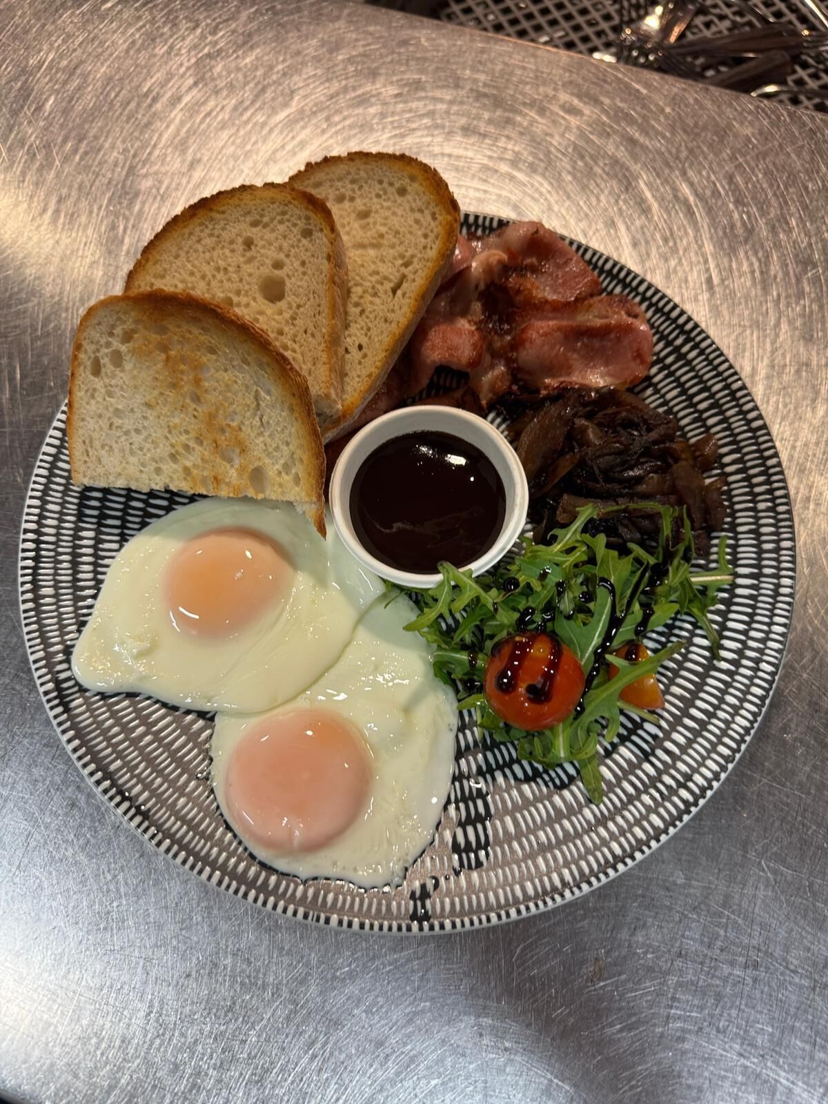 A plate with toast, bacon, eggs, a salad, and a small bowl of sauce.