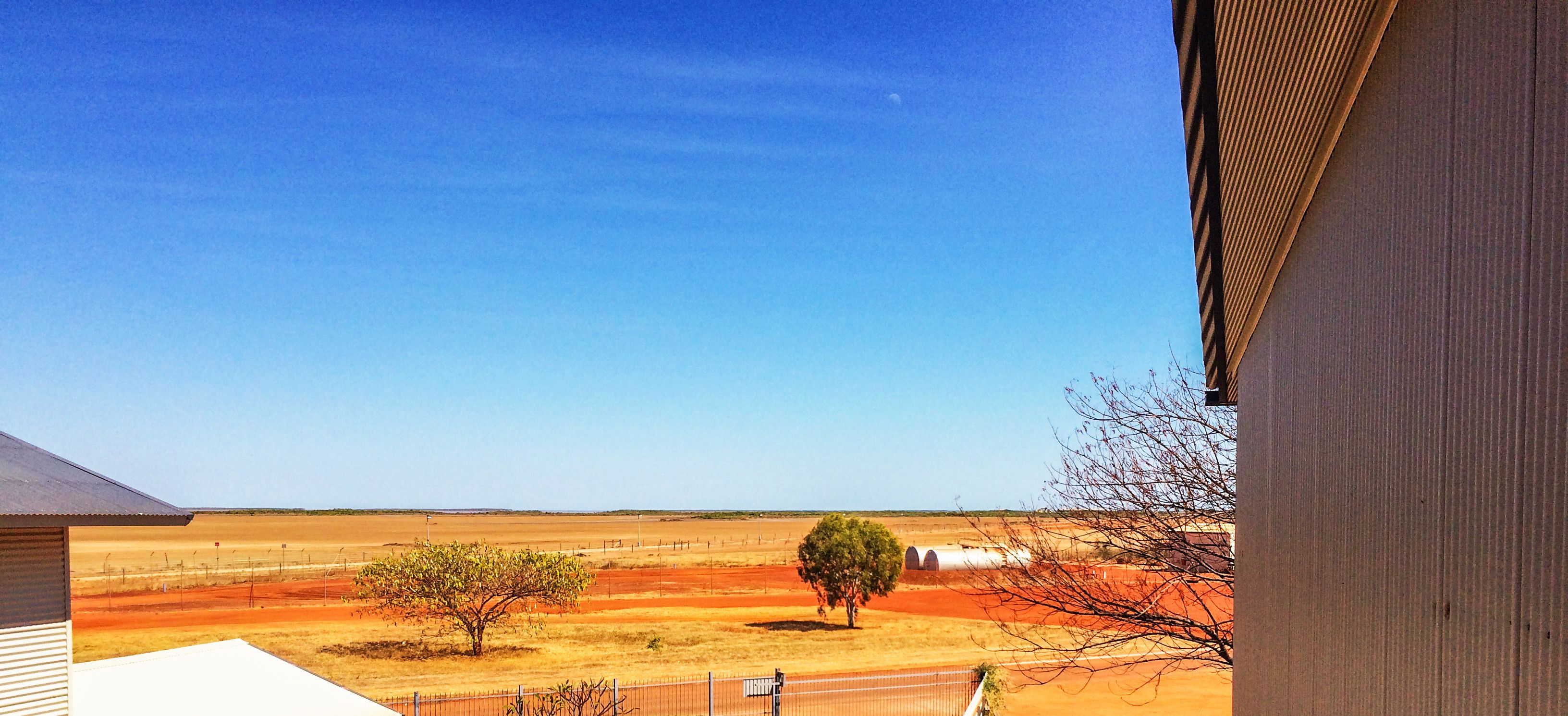A rural landscape with a clear blue sky, a fenced area with sparse vegetation, and a distant building.