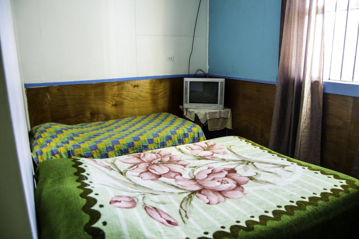 Um quarto pequeno com duas camas, uma com colcha florida e outra com padrão xadrez. Há uma pequena mesa com uma televisão em cima.