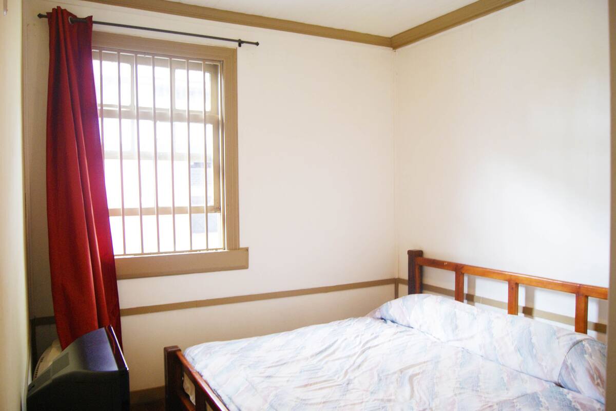 Quarto com uma cama de solteiro, janela com persianas e cortina vermelha.
