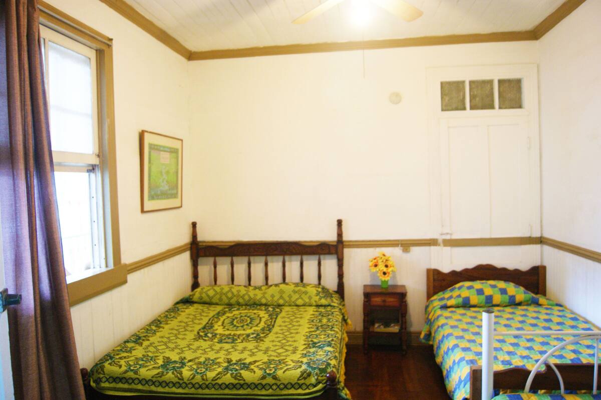 Quarto com duas camas, cabeceira de madeira, mesa de cabeceira pequena com um vaso e janela com cortinas.