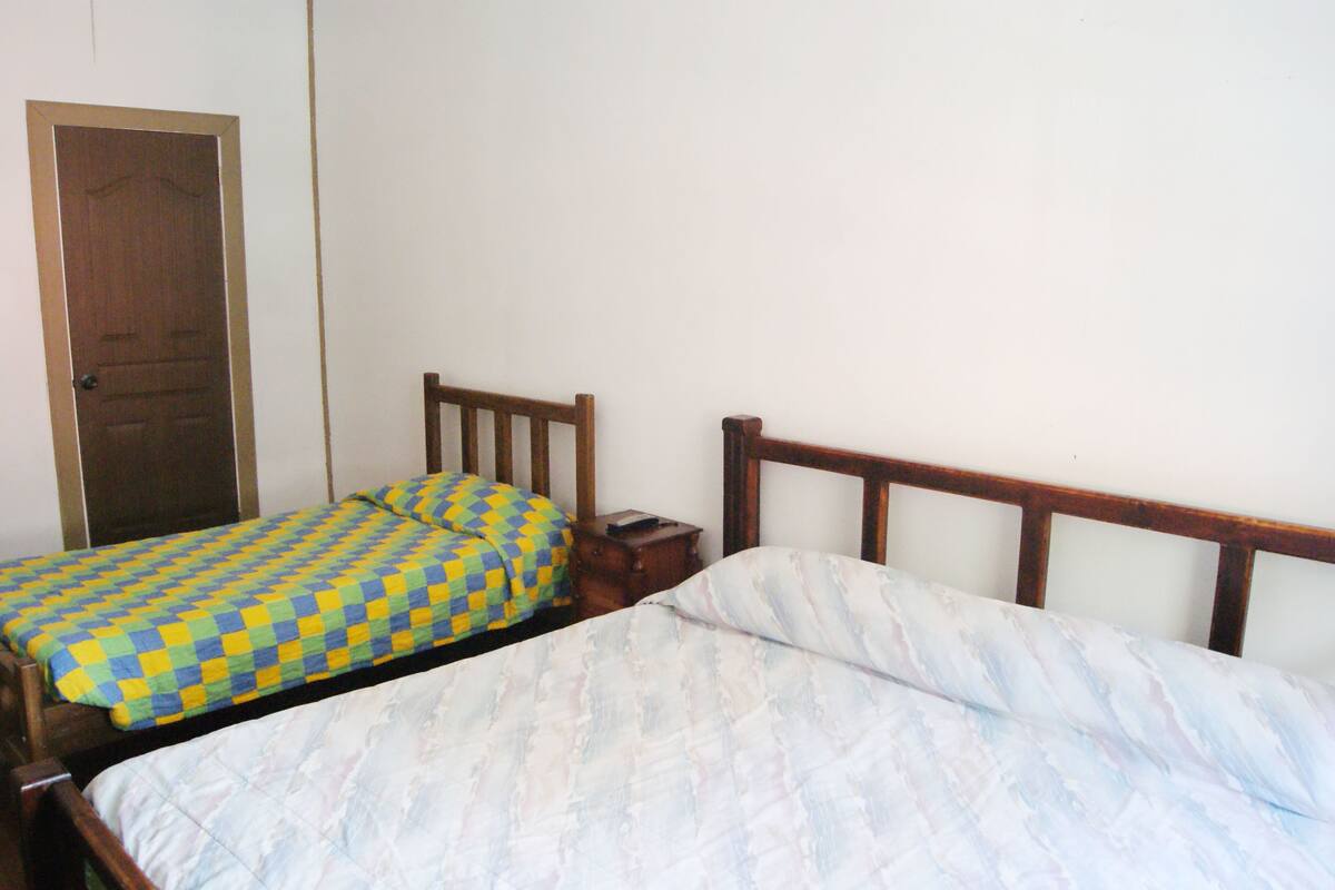 Quarto com duas camas de solteiro, cabeceiras e mesa de cabeceira de madeira, e uma porta.