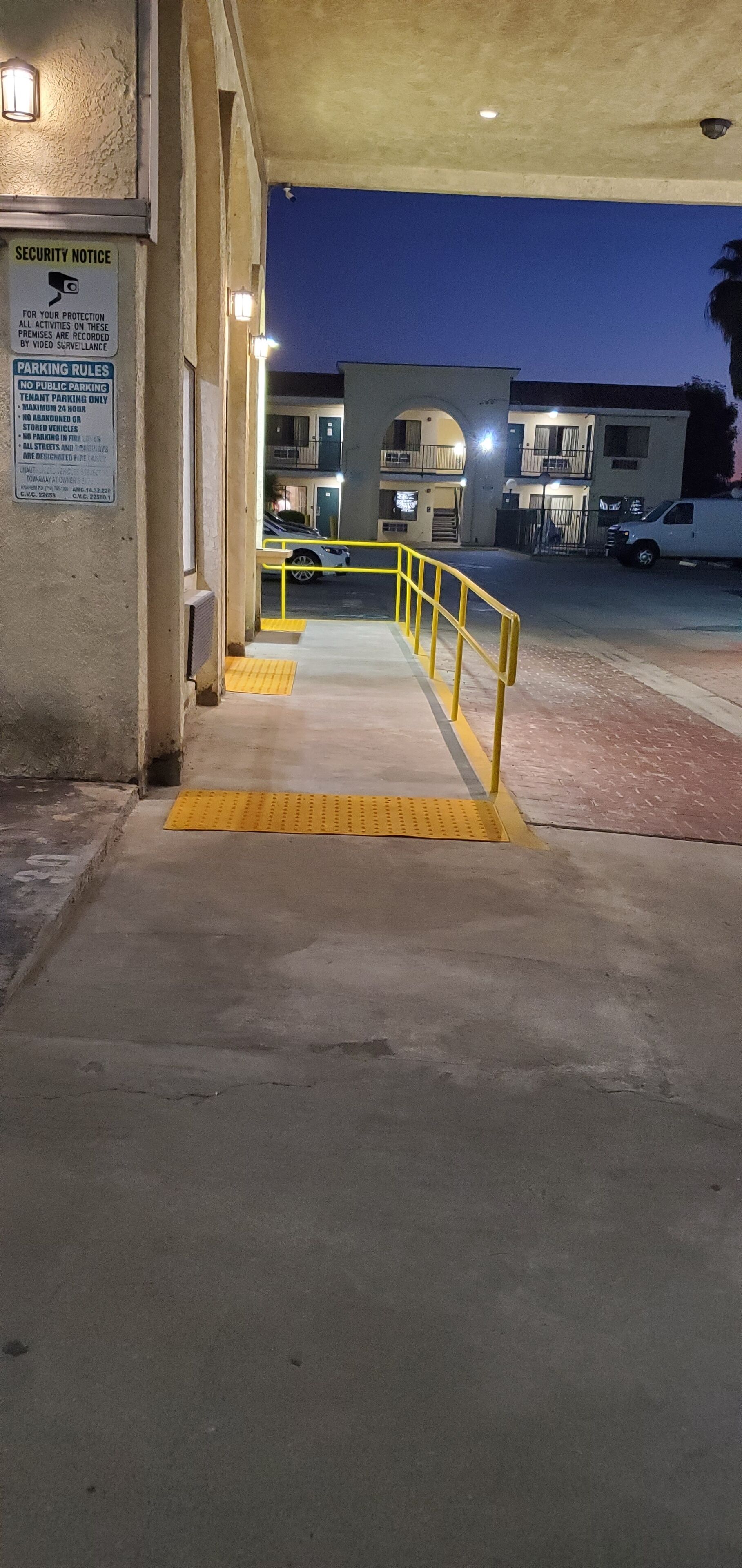 A nighttime view of a building entrance with a security notice, parking rules, and a yellow safety ramp leading to a gated parking area.