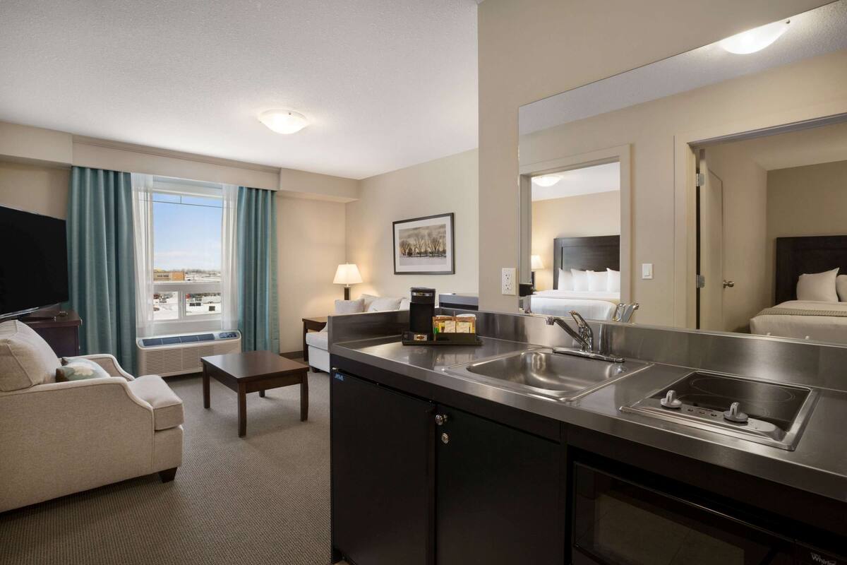 A hotel room with a kitchenette, a sofa, a coffee maker, and a large window with curtains.