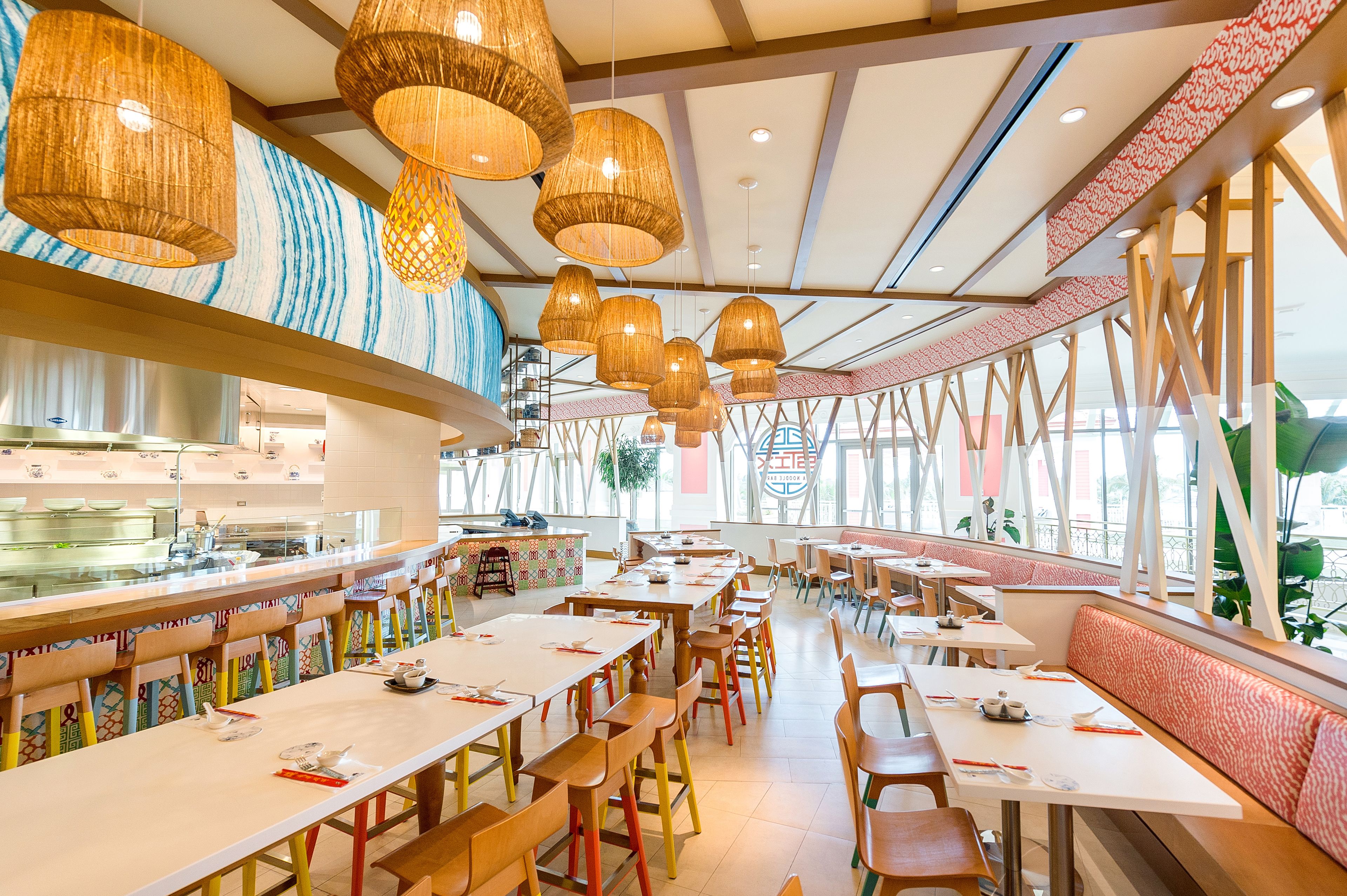 Un restaurante espacioso con techos altos, candelabros de mimbre colgantes y una larga barra, con vislumbre de una cocina al fondo.
