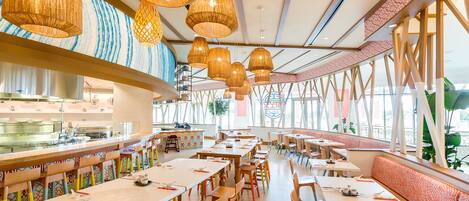 Un restaurante espacioso con techos altos, candelabros de mimbre colgantes y una larga barra, con vislumbre de una cocina al fondo.
