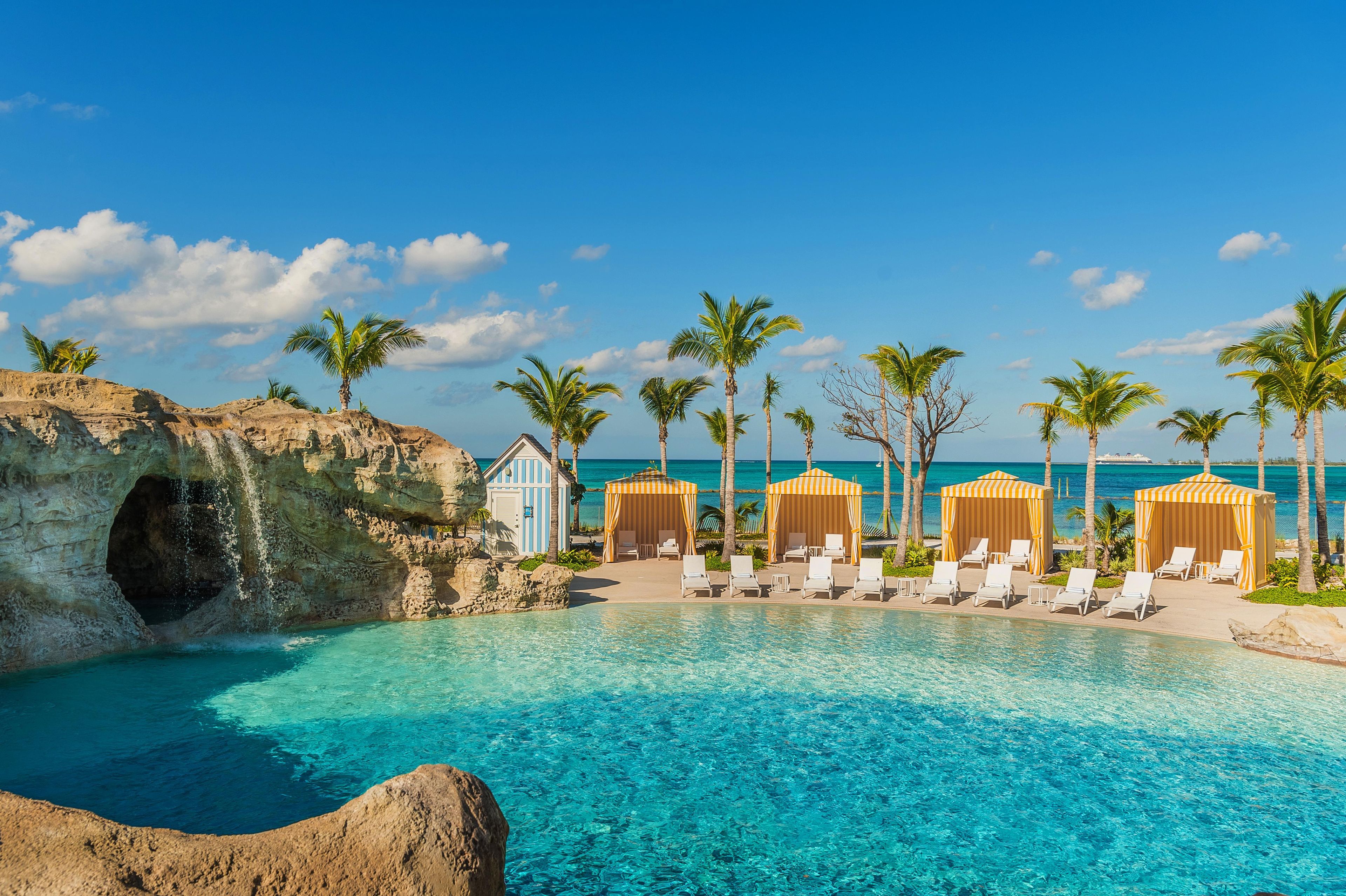 Zona de piscina con agua azul cristalina, palmeras y cabañas de playa.