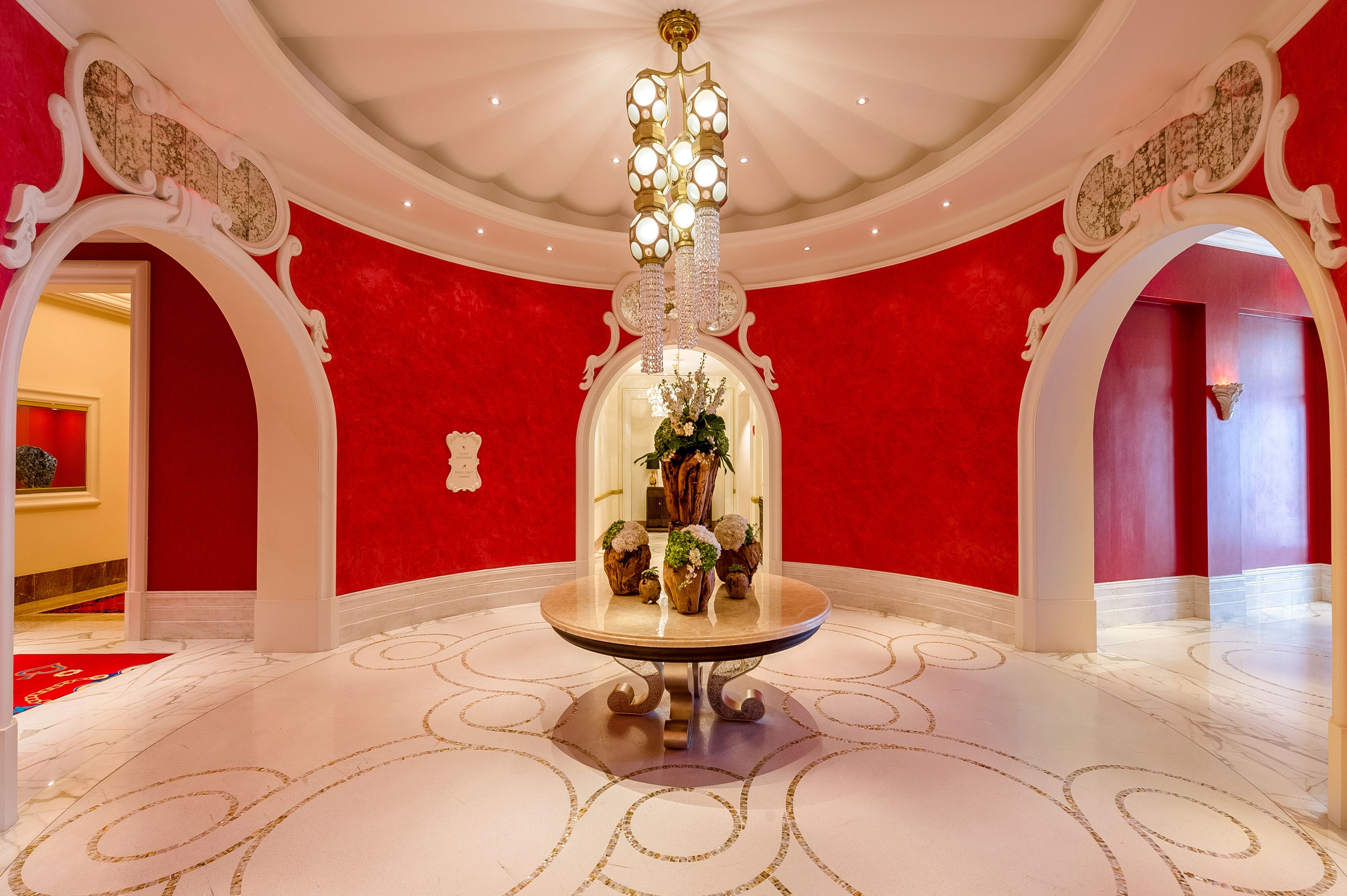 Un vestíbulo imponente con una pared roja, suelo de mármol blanco y una mesa central con candelabro.