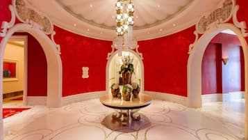 Un vestíbulo imponente con una pared roja, suelo de mármol blanco y una mesa central con candelabro.