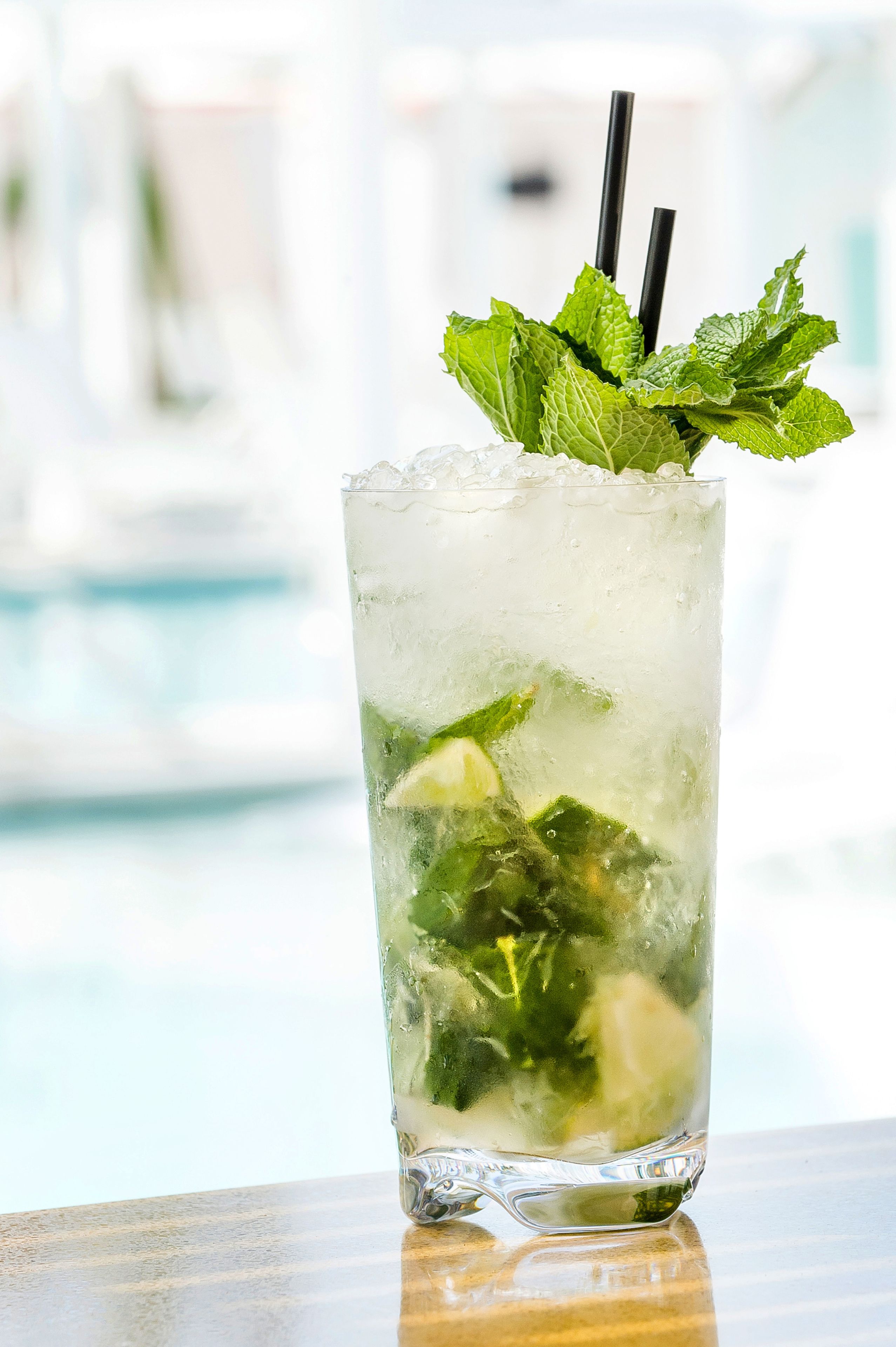 A tall glass with ice, mint leaves, and lime slices, with black straws, on a wooden surface.