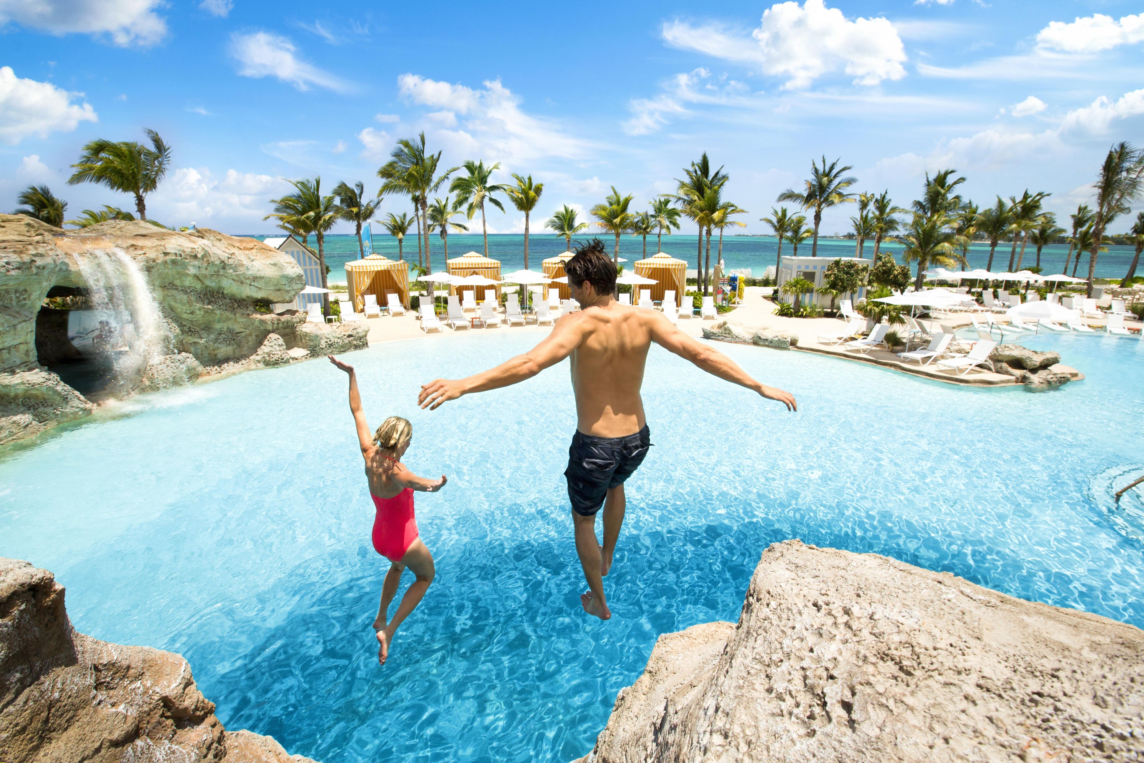 A man and a woman jumping into a pool with a tropical resort in the background.