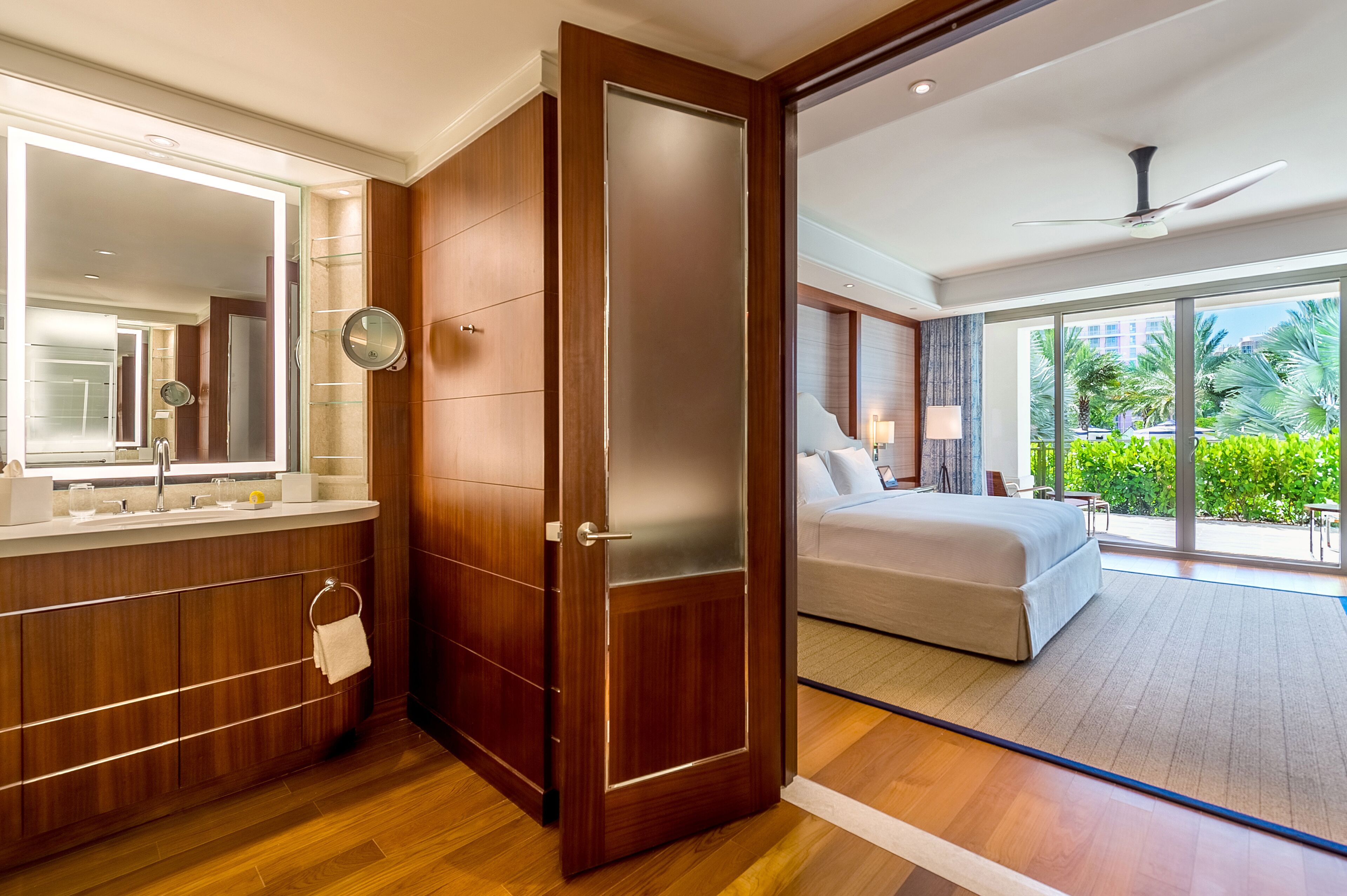 Une chambre d’hôtel moderne dotée d’une coiffeuse en bois, d’un grand miroir, d’un lit donnant sur l’extérieur et d’un ventilateur de plafond.