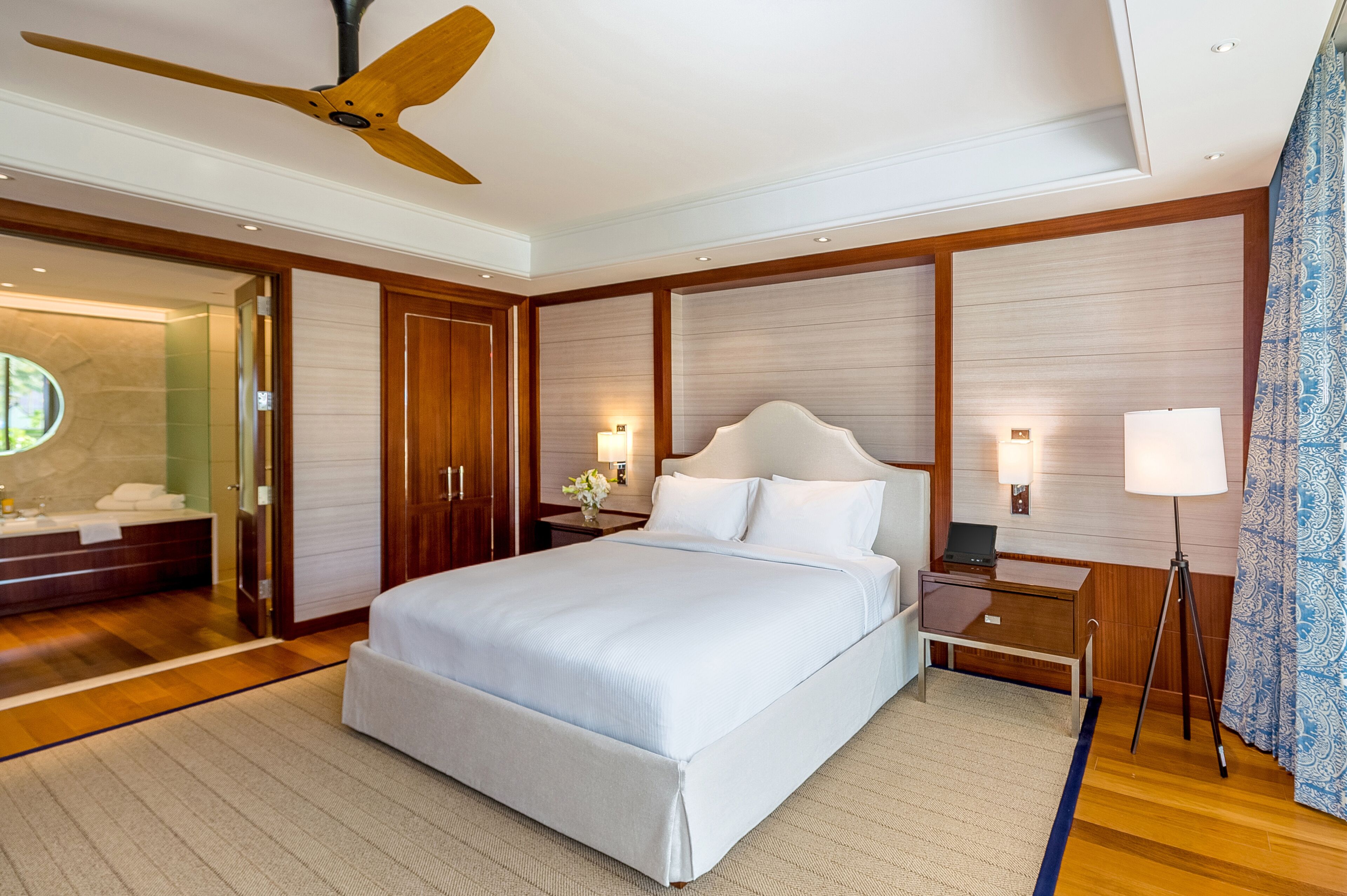 Un'ampia camera d'hotel con un grande letto, pannelli in legno, una ventola a soffitto, un comò con specchio e un comodino con lampada.
