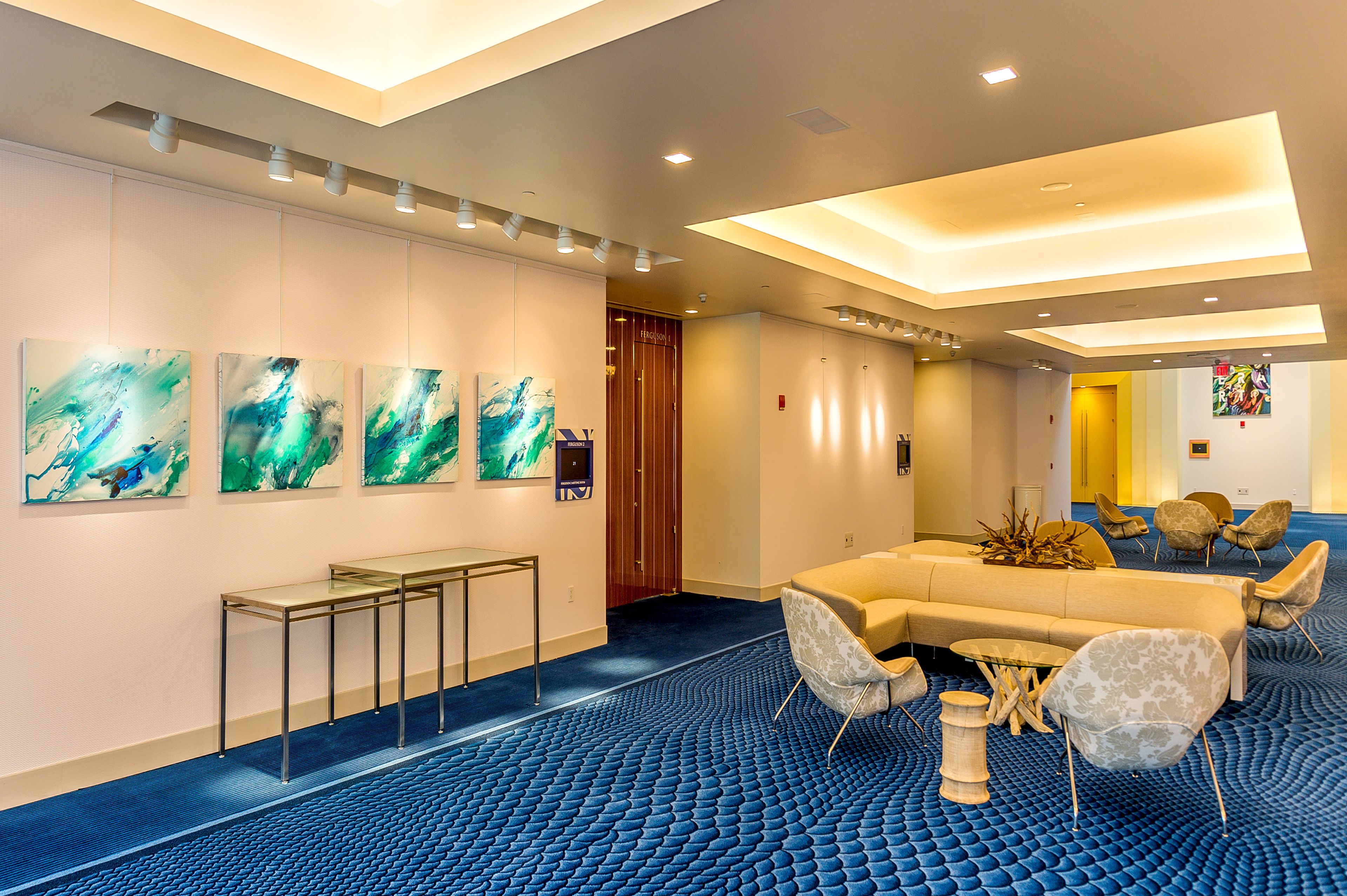 A hotel lobby with a blue carpet, a sofa, chairs, a table, and abstract paintings on the wall.