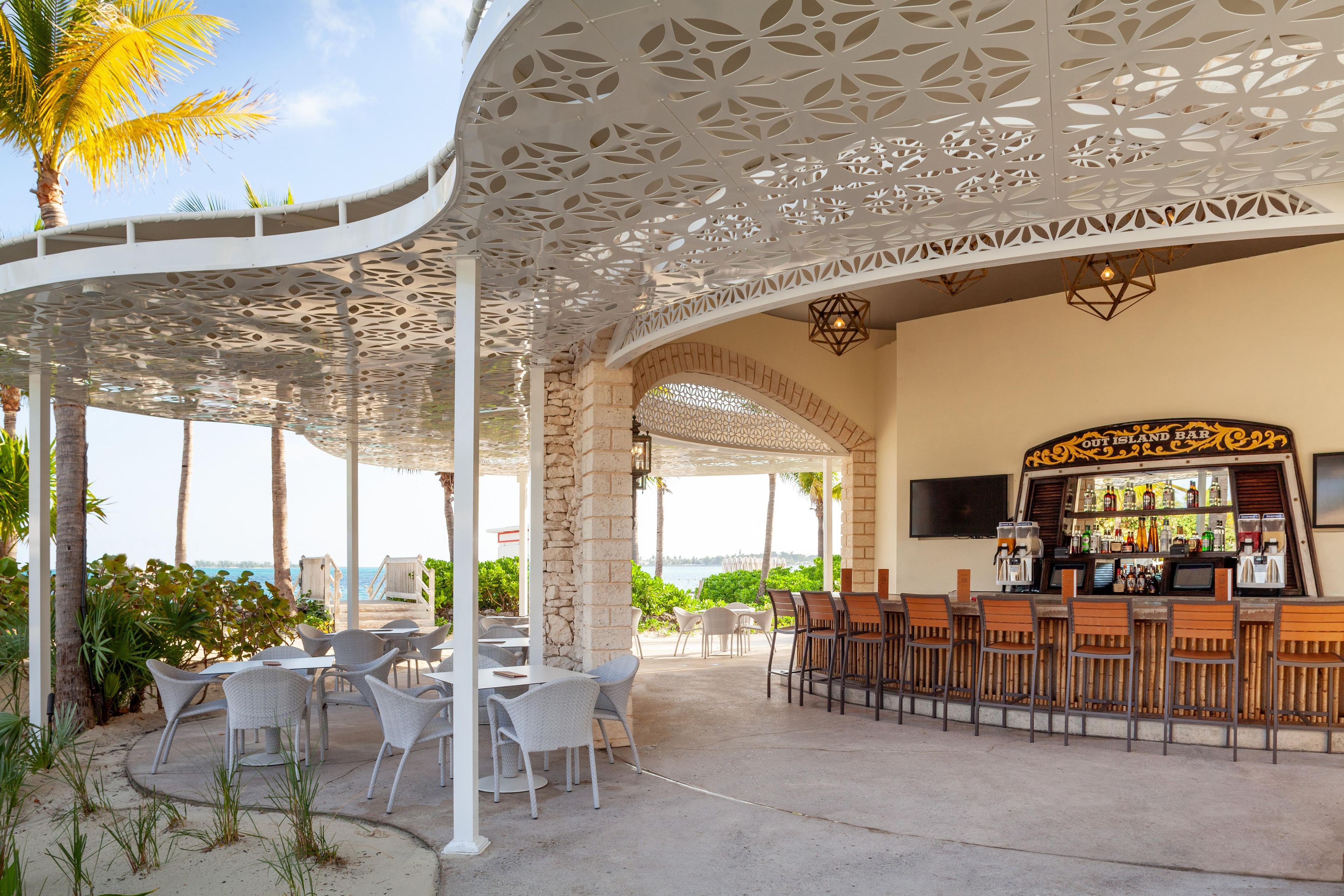 Zona de bar al aire libre con vista al mar, que presenta un techo con diseño, asientos y una barra con un letrero que dice 'OUT ISLAND BAR'.
