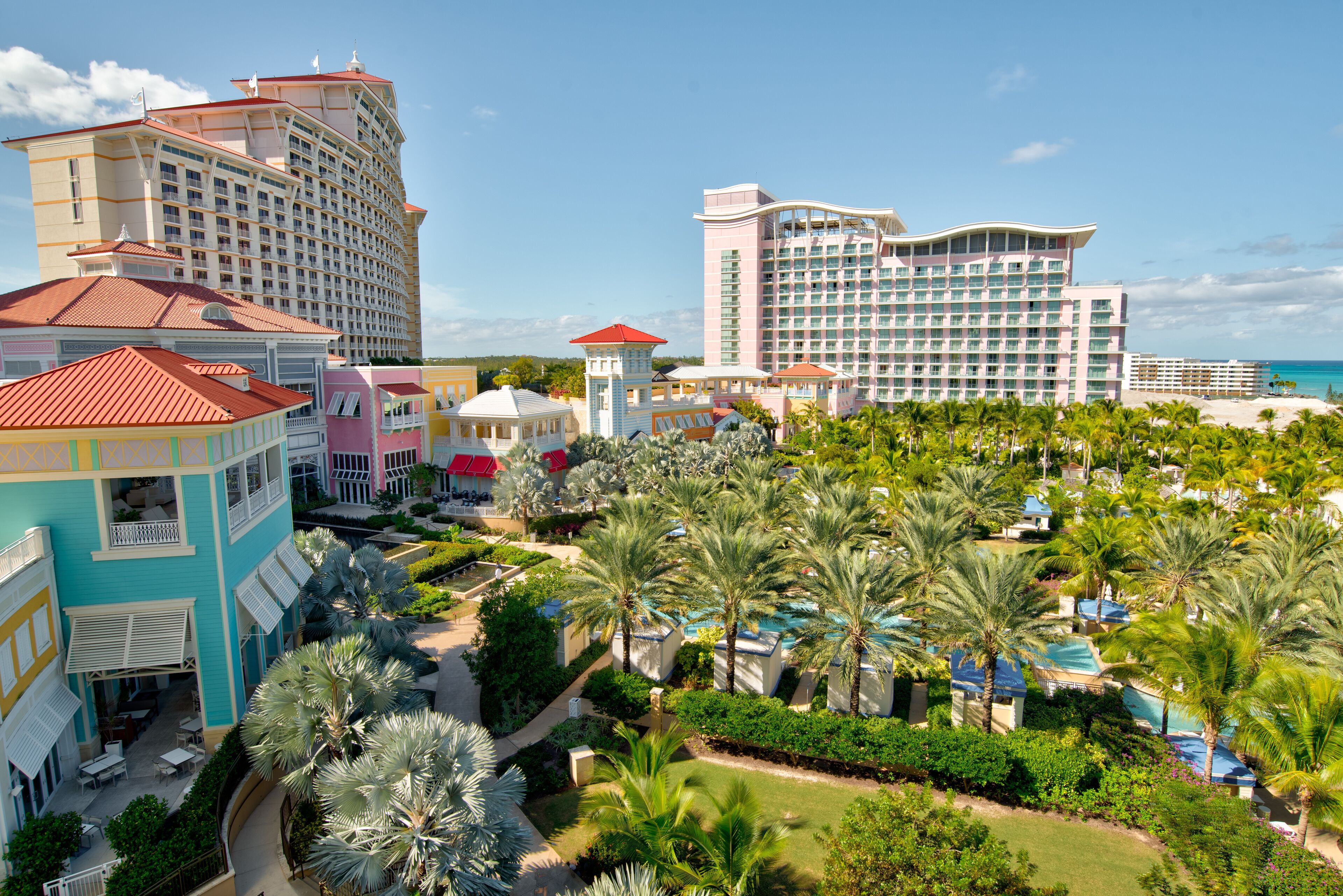Een resort met meerdere gebouwen, waaronder een groot hotel met een gebogen dak, een kleiner gebouw met een rood dak en een toren met een rood dak. Er staan palmbomen en er is een zwembad te zien.