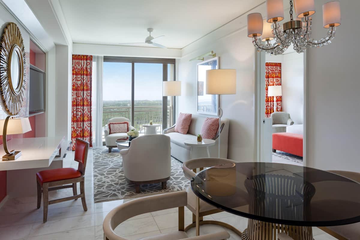 Habitación de hotel con una mesa de comedor, un sofá, una silla, un televisor y un ventanal grande con cortinas.