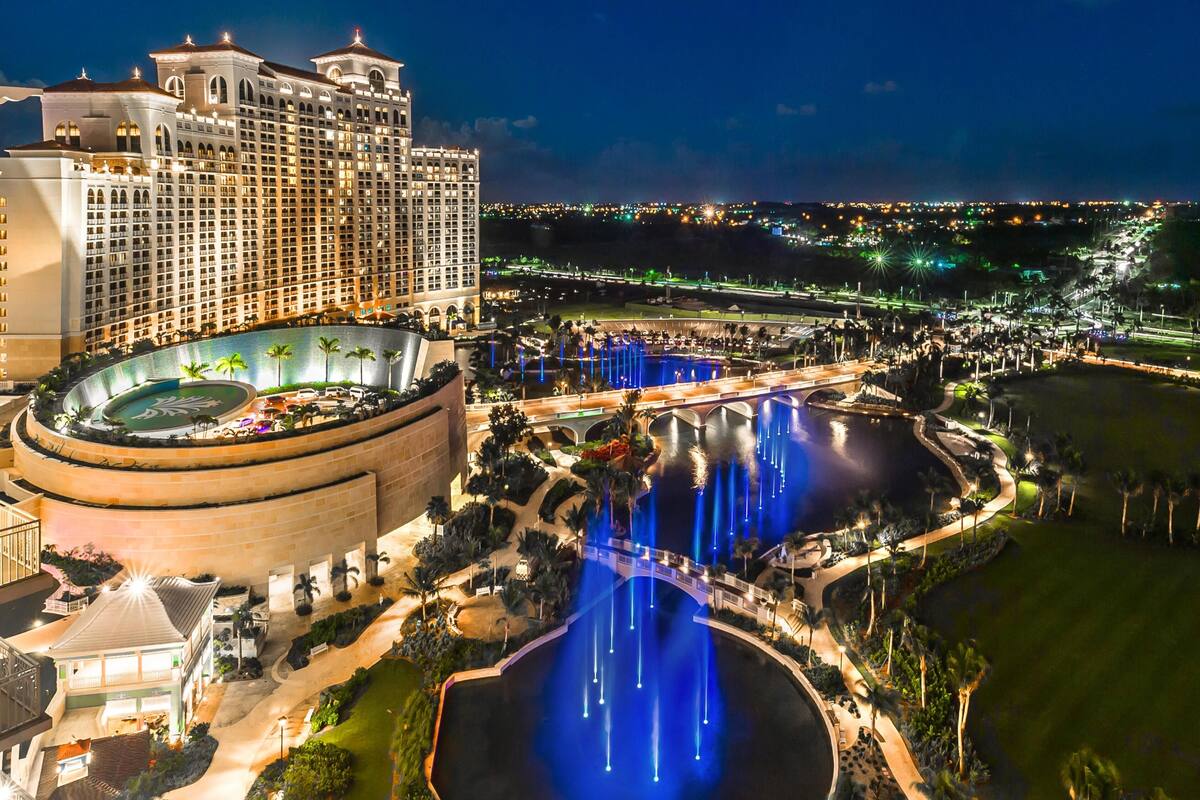Un hotel grande con zona de piscina que incluye fuentes de agua, rodeado de palmeras y un paisaje bien iluminado.