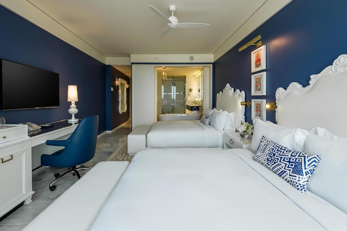 Una habitación de hotel con escritorio, dos camas, una silla, ventilador de techo y televisión.
