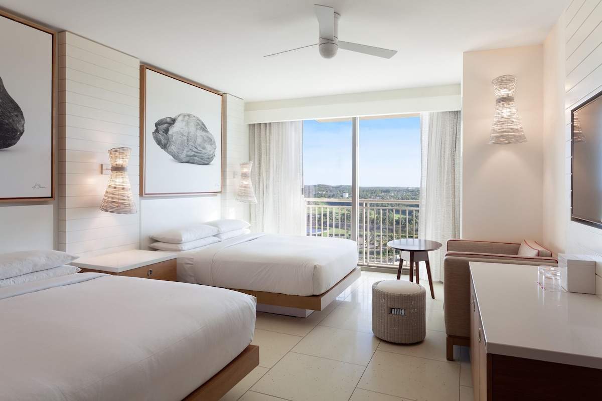 A hotel room with two beds, a balcony with a city view, a desk, and a ceiling fan.