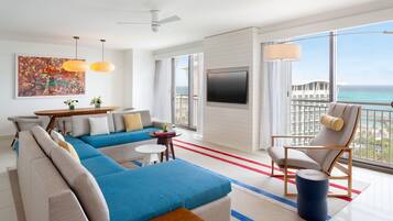 A modern living room with a large sofa, a dining area, a TV mounted on the wall, and a balcony with a view of the ocean.