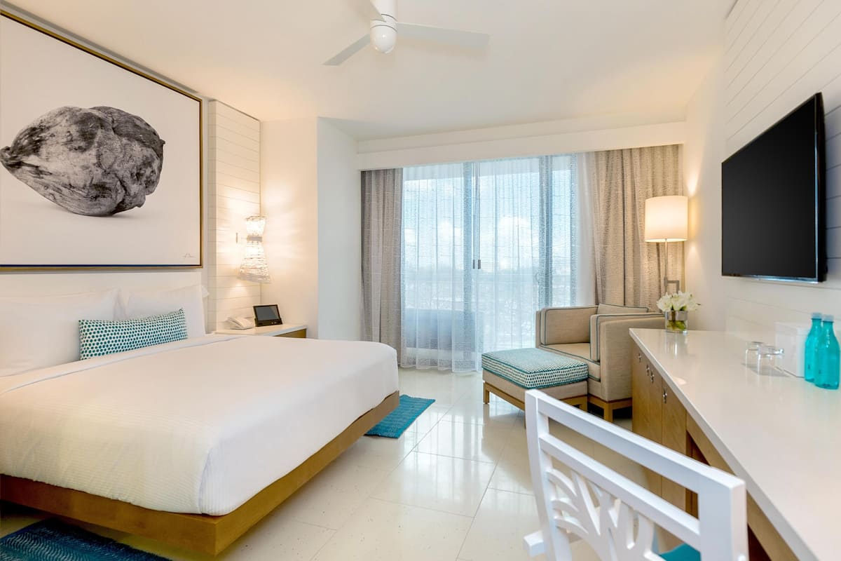 Habitación de hotel con cama, escritorio, silla, televisión y ventanas grandes.