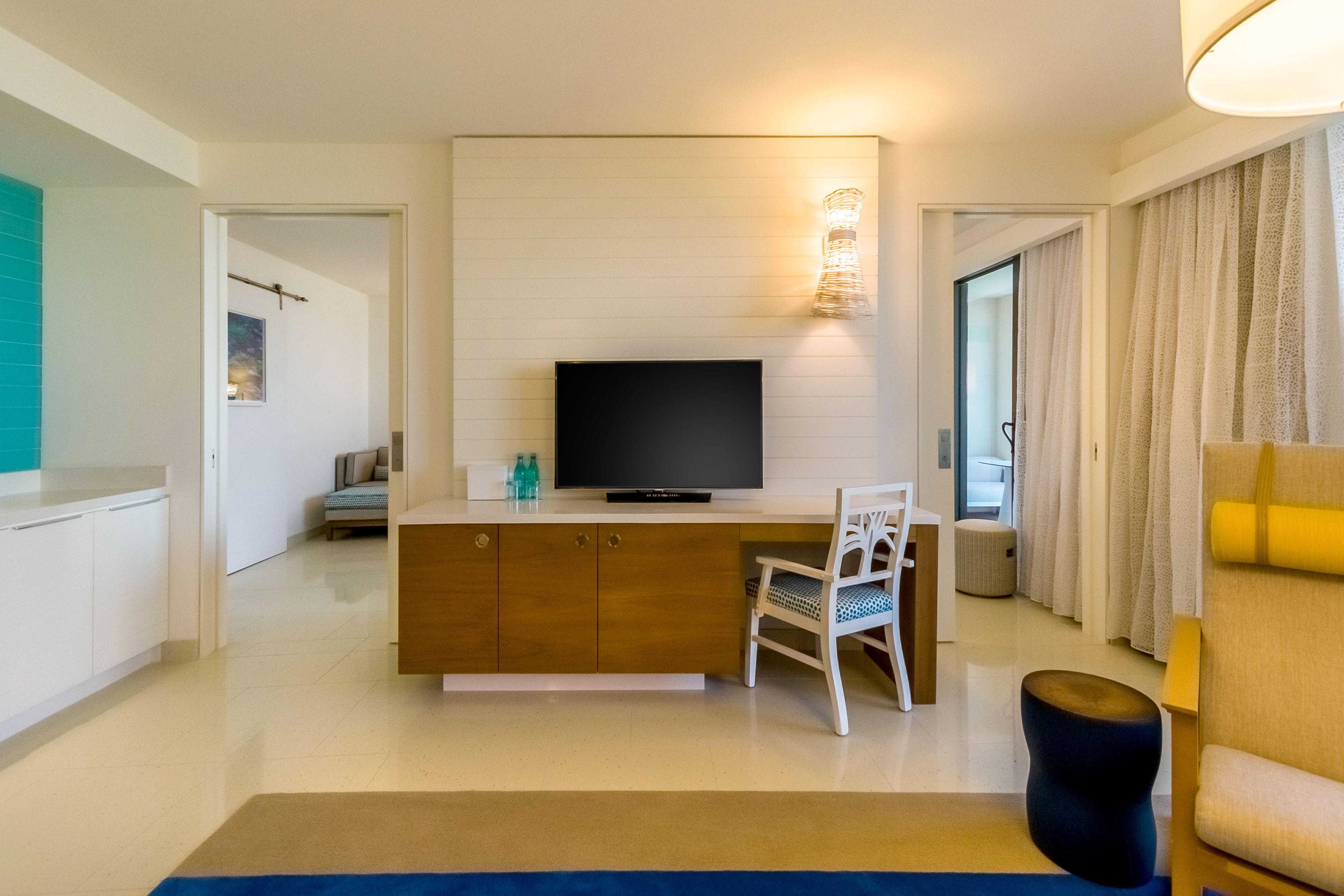 A modern hotel room with a flat-screen TV on a wooden cabinet, a white chair, a small round ottoman, and a sofa with a yellow cushion.