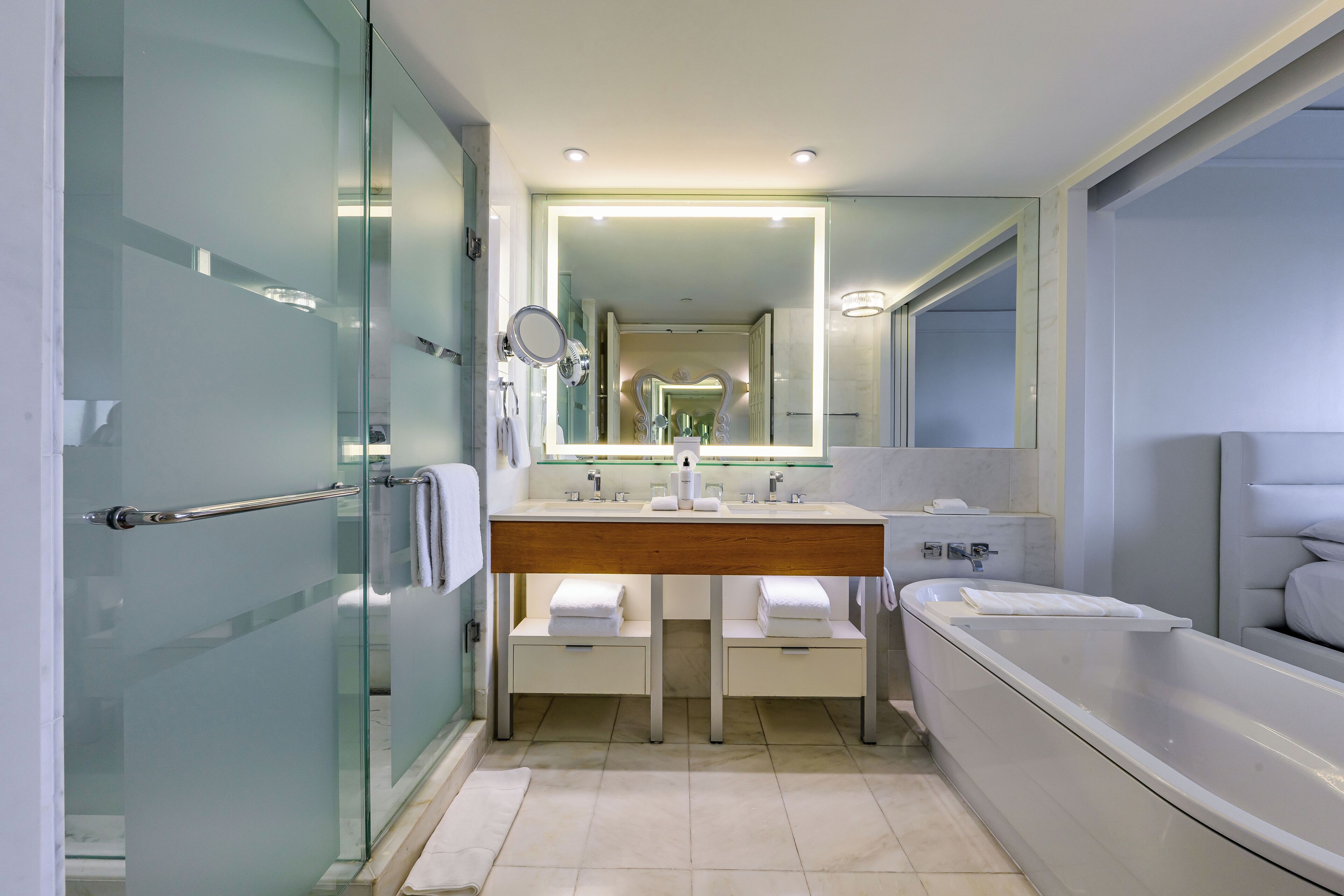 Ein modernes Badezimmer mit einer Duschkabine aus Glas, einem großen Spiegel, einem Holzwaschtisch, einer Badewanne und einem weiß gefliesten Boden.
