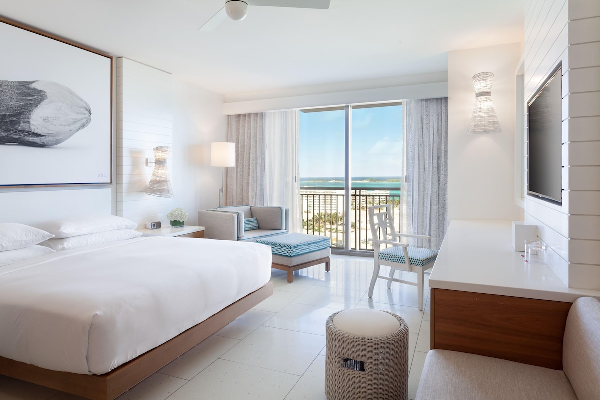A hotel room with a bed, a desk, a chair, a small table, a waste basket, and a large window with a view.
