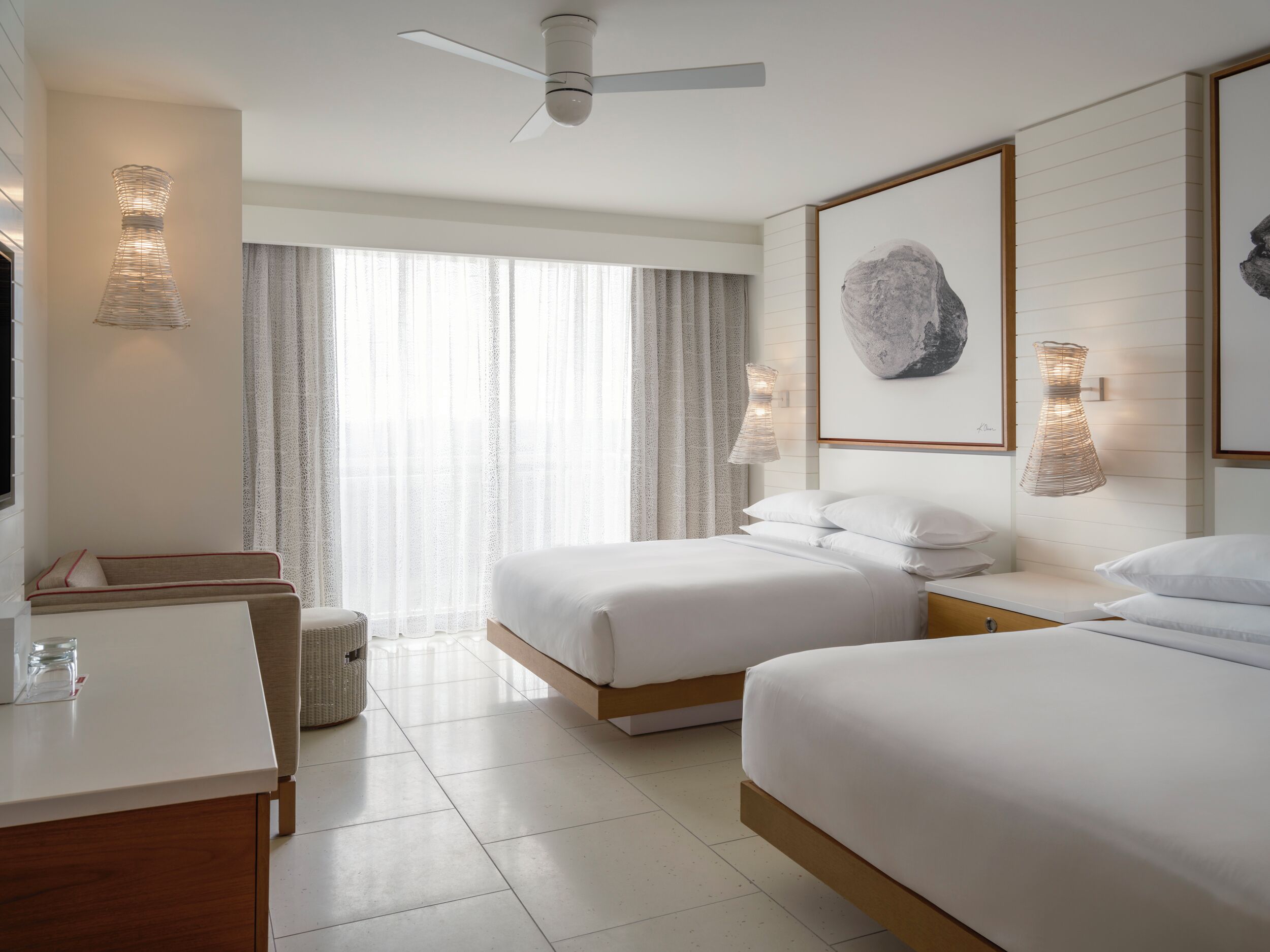 Habitación de hotel con dos camas, un escritorio, una silla, un ventilador de techo y un ventanal grande con cortinas.