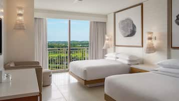 A hotel room with two beds, a desk, a chair, a balcony with a view, and a ceiling fan.