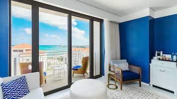 A hotel room with a large sliding glass door leading to a balcony with a view of the ocean and buildings. There is a white sofa with a patterned pillow, a wooden chair, and a small white table with a tray of items.