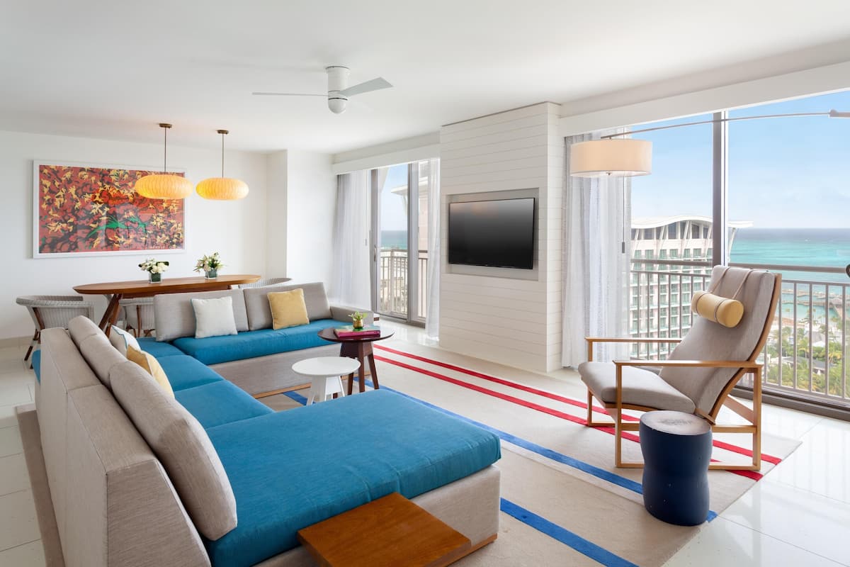 Una habitación de hotel moderna con un balcón grande, un televisor de pantalla plana y una zona de estar con un sofá y sillones.