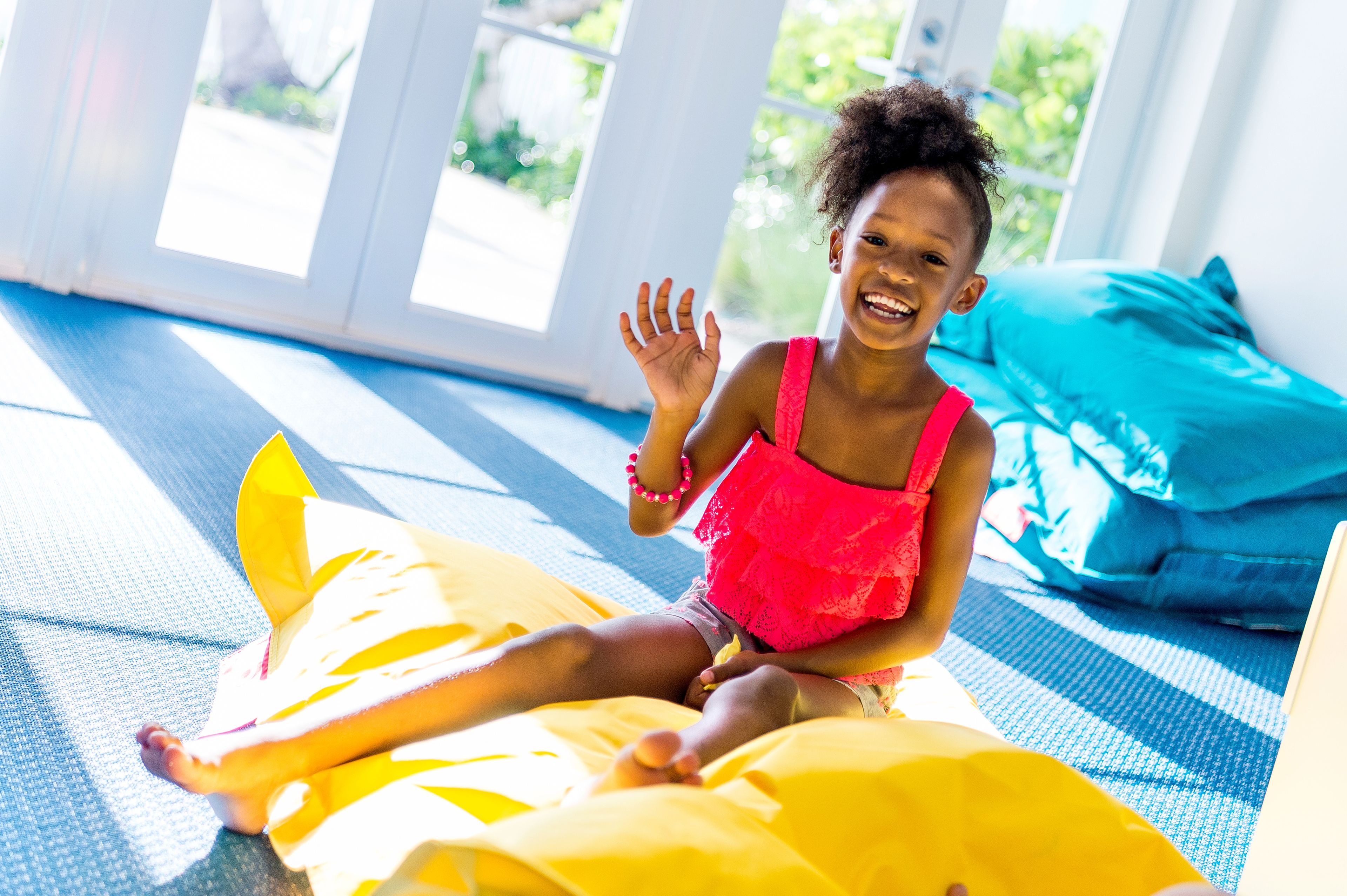 Un enfant est assis sur un pouf jaune dans une pièce dotée de grandes fenêtres et d’un tapis bleu.