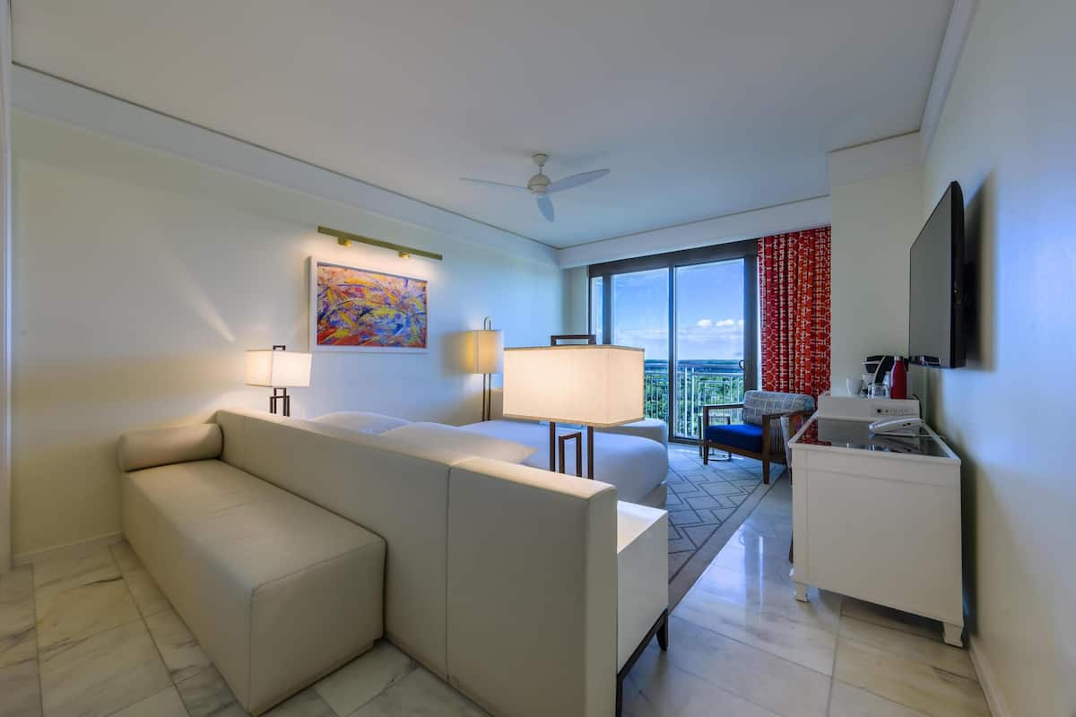 Habitación de hotel con sofá, escritorio con lámpara, televisor, balcón con cortina roja y un cuadro en la pared.