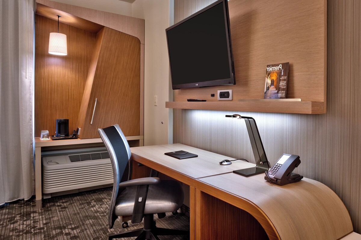 Une chambre d’hôtel équipée d’un bureau, d’une chaise, d’une télévision et d’une petite étagère sur laquelle se trouve un livre.