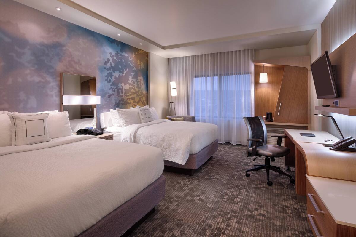 A hotel room with two beds, a desk, a chair, a flat-screen TV, and a large window with curtains.