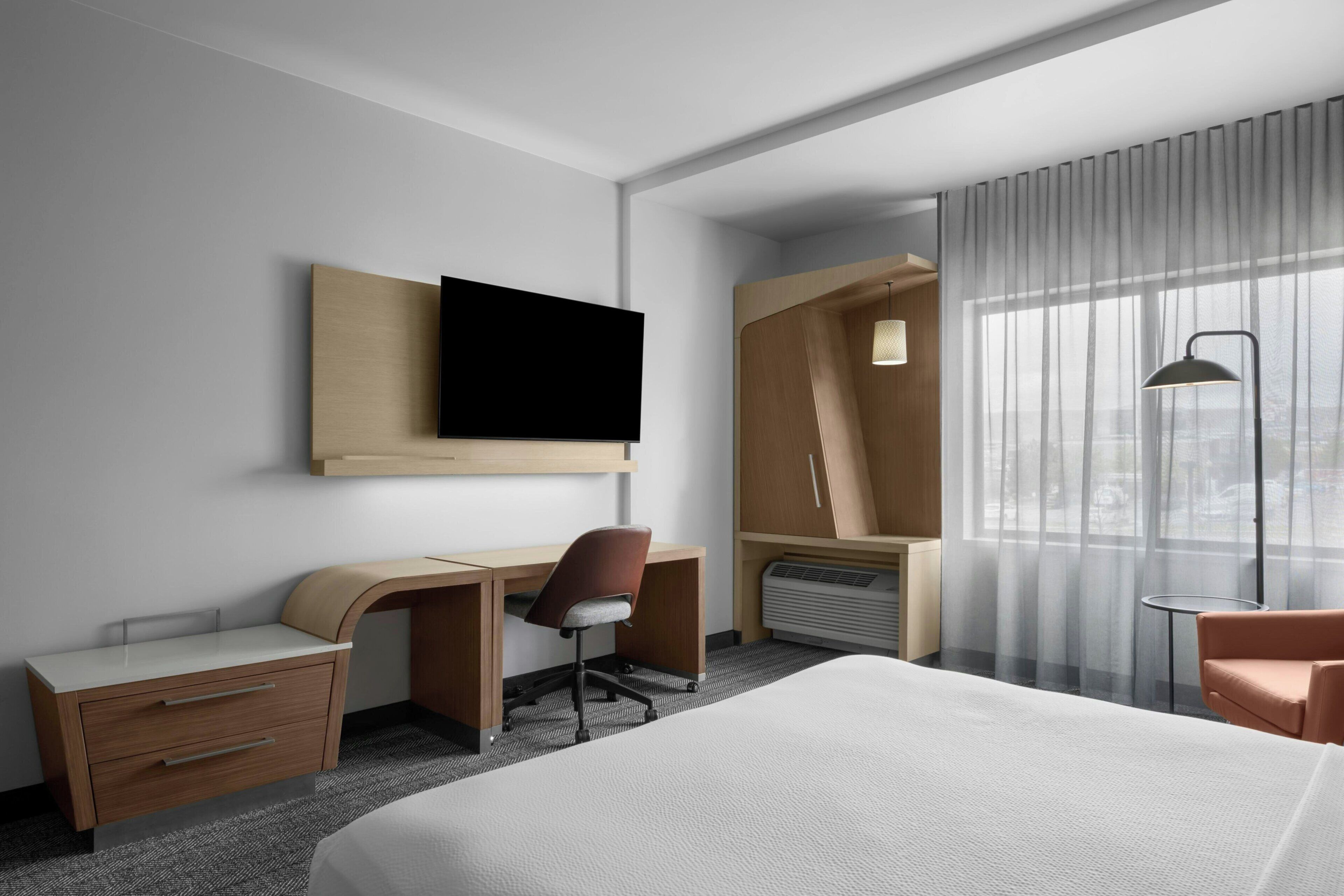 Ett hotellrum med en säng, ett skrivbord, en stol, en tv, en värmare och ett fönster med gardiner.