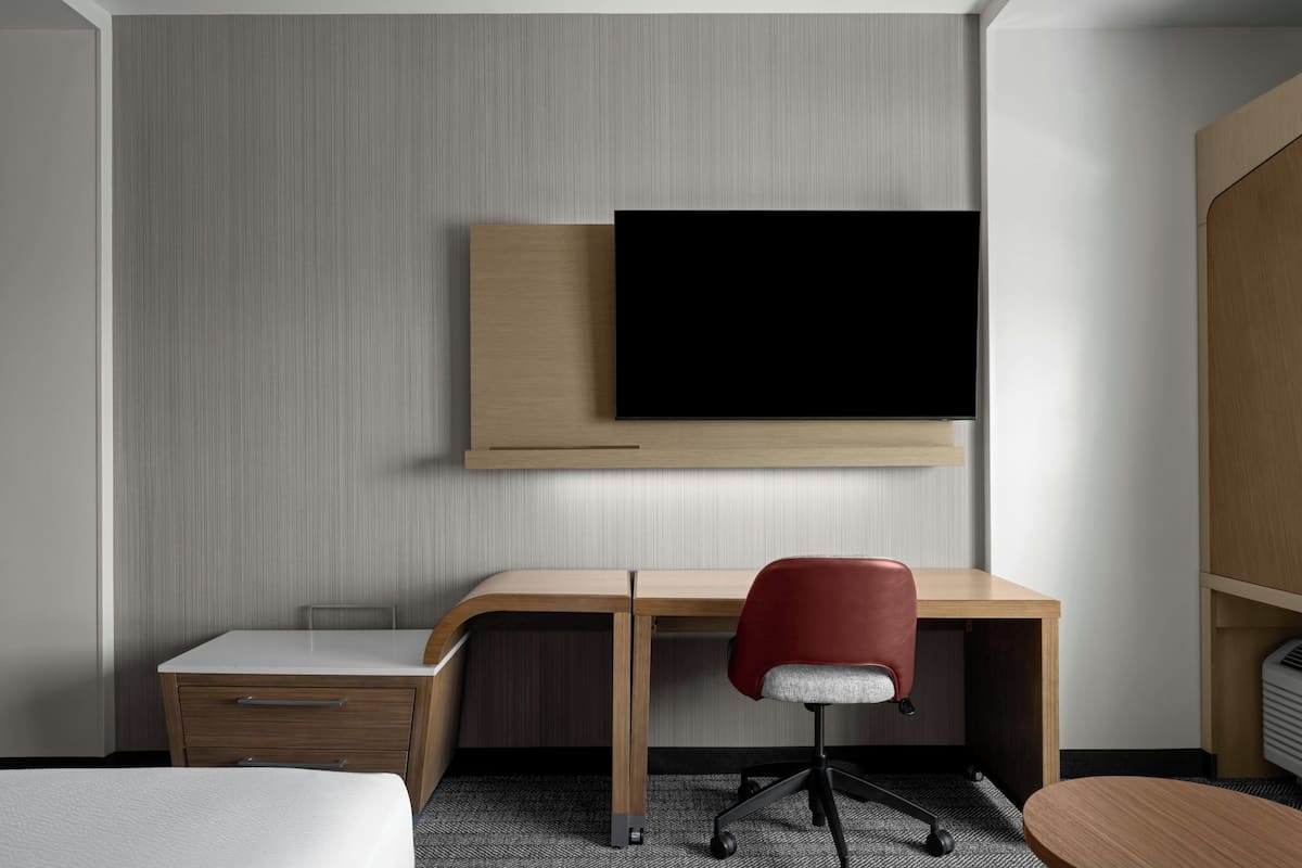 Una habitación de hotel con un escritorio de madera, una silla de oficina roja, un televisor de pantalla plana montado en la pared, una cama y una mesita de noche pequeña.