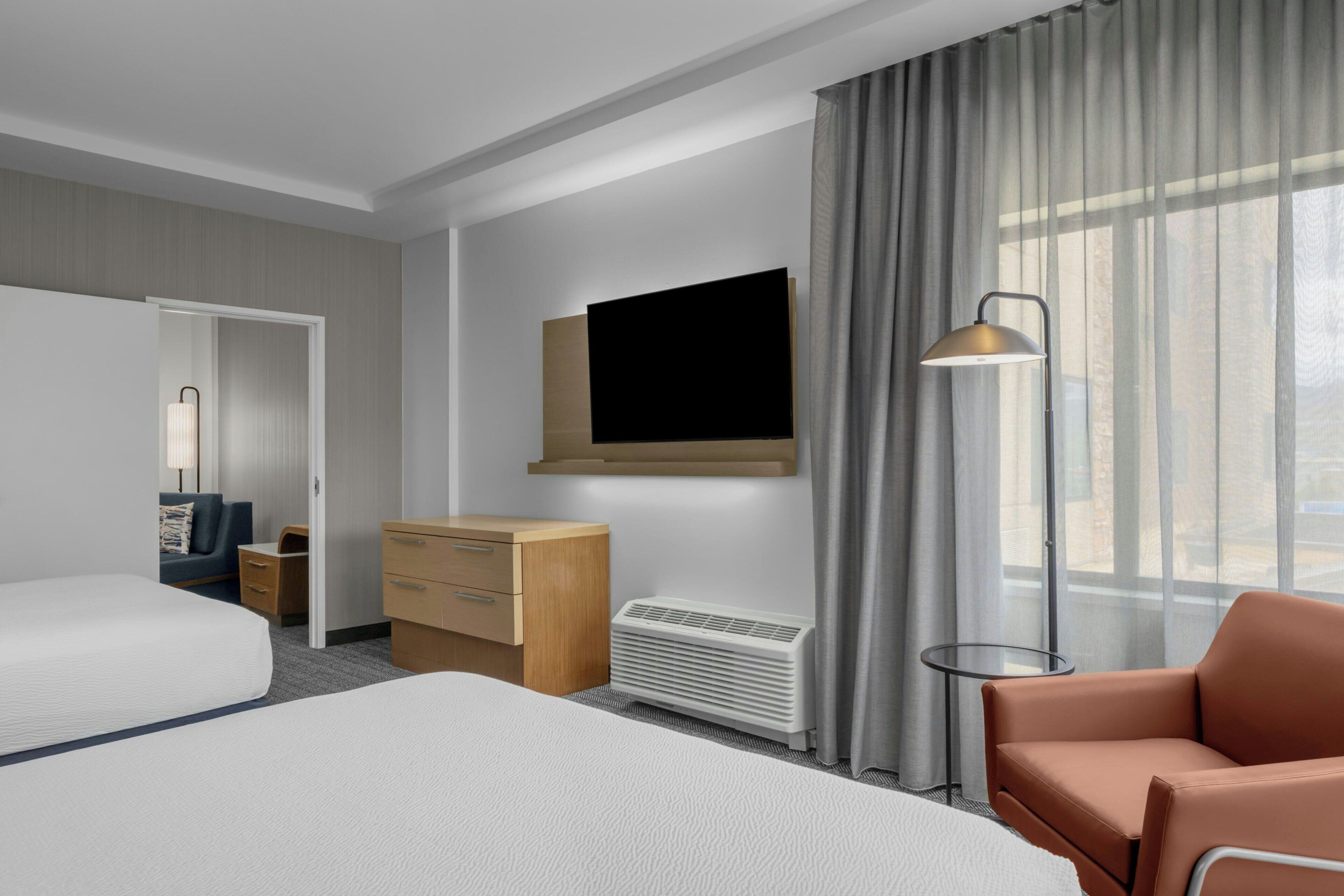 Quarto de hotel com cama, escrivaninha, cômoda, televisão, abajur e janela com cortinas.
