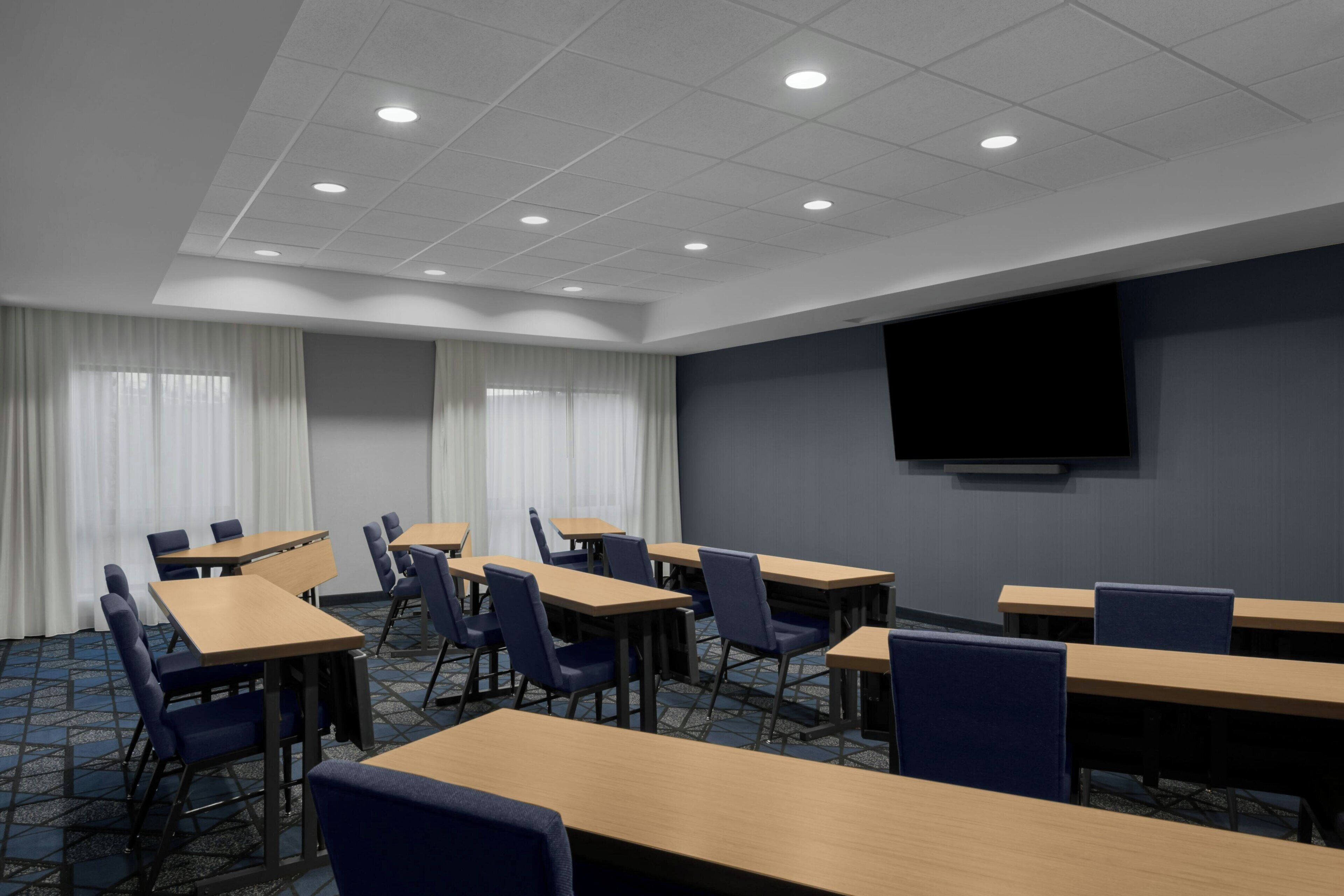 A conference room with multiple tables and chairs, a large screen on the wall, and a ceiling with recessed lighting.