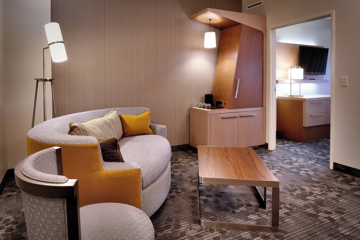 A modern hotel room with a curved sofa, a small wooden table, a desk with a lamp, and a flat-screen TV mounted on the wall.