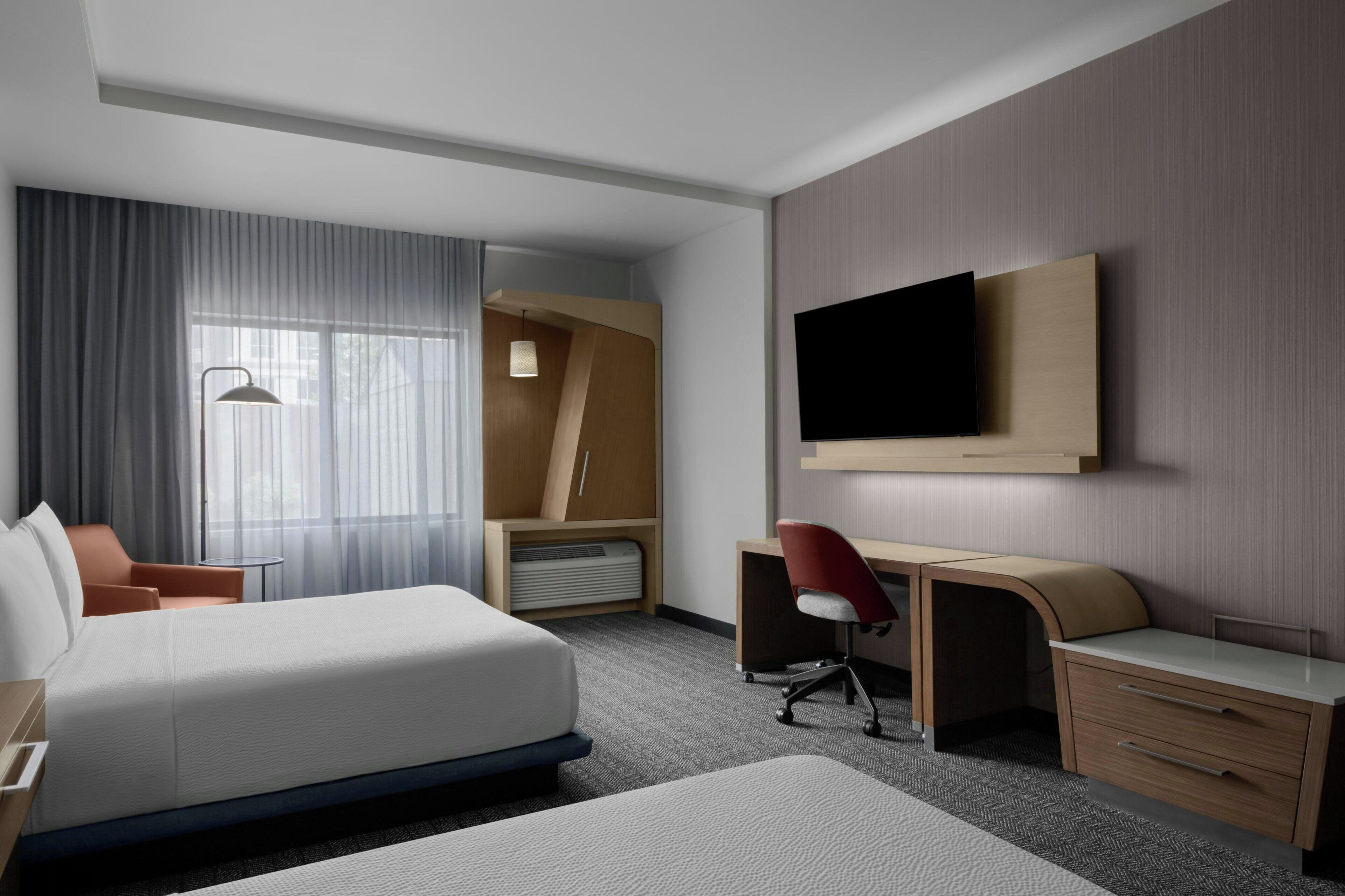 Quarto de hotel com cama, escrivaninha, cadeira, televisão e uma janela grande com cortinas.