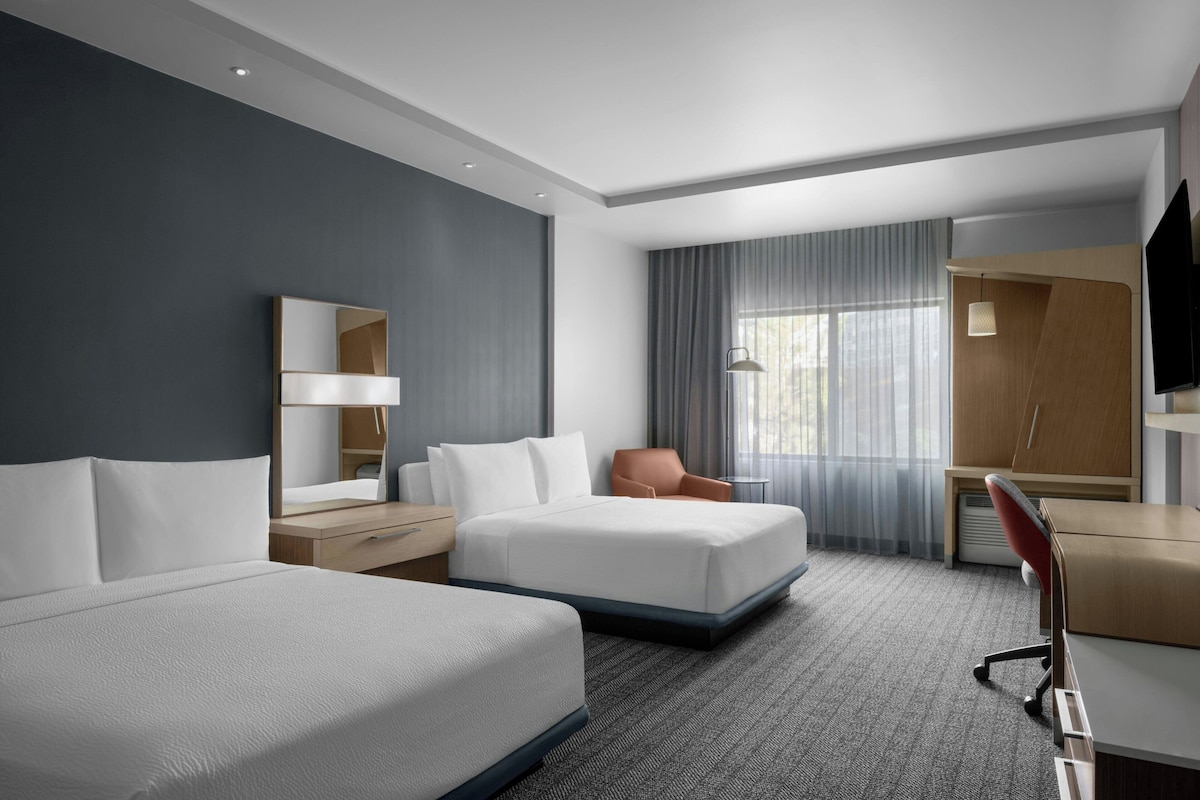 A hotel room with two beds, a desk, a chair, a TV, and a large window with curtains.