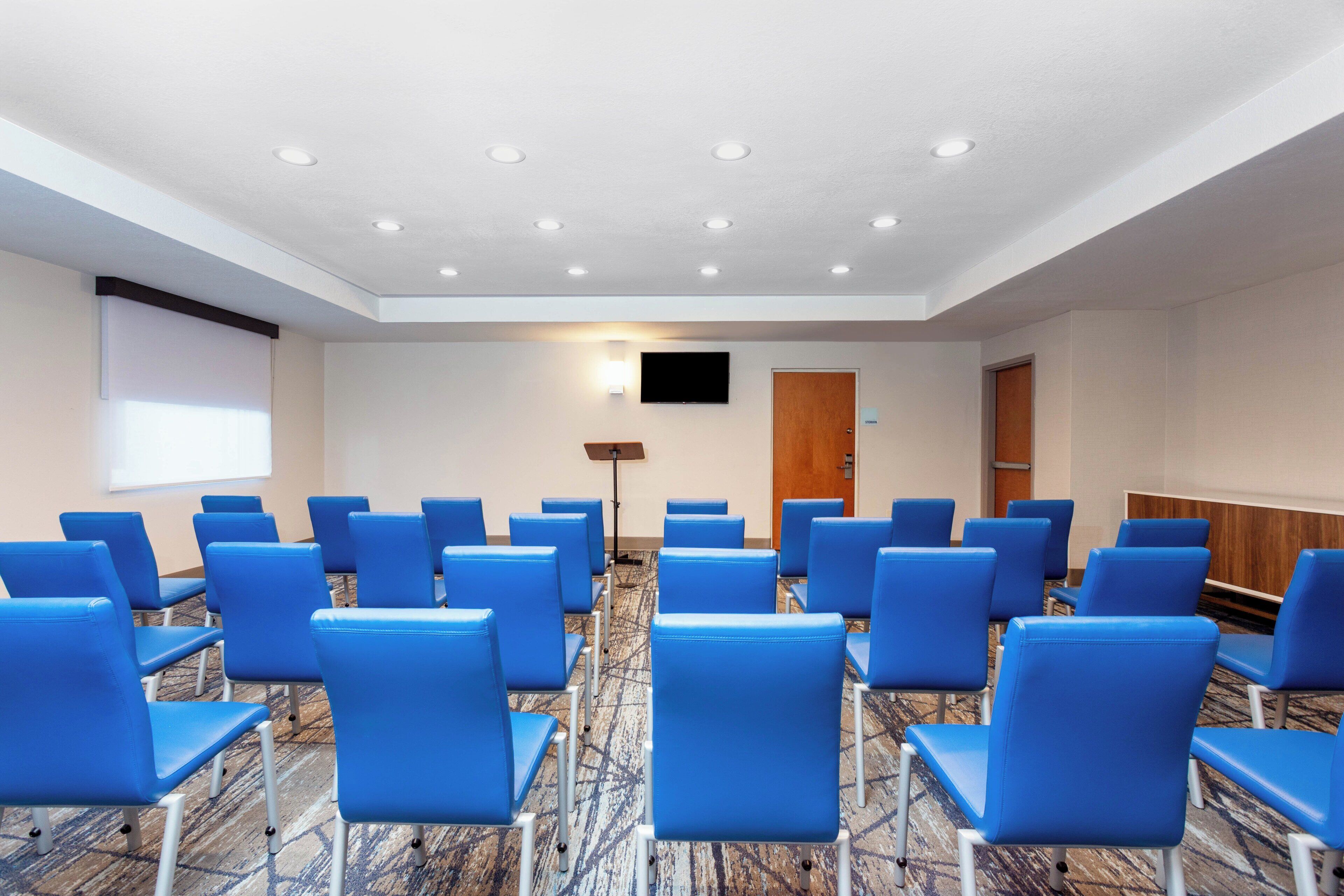 Ein Konferenzraum mit blauen Sitzreihen, einer Projektionsleinwand, einem Flachbildfernseher und einem Teppichboden.