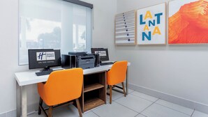 A hotel reception desk with two computer monitors, a printer, and two orange chairs. There are framed artworks on the wall and a window with blinds.