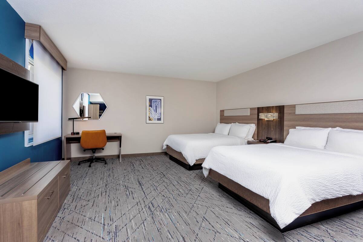 A hotel room with two beds, a desk, a chair, a TV, and artwork on the walls.