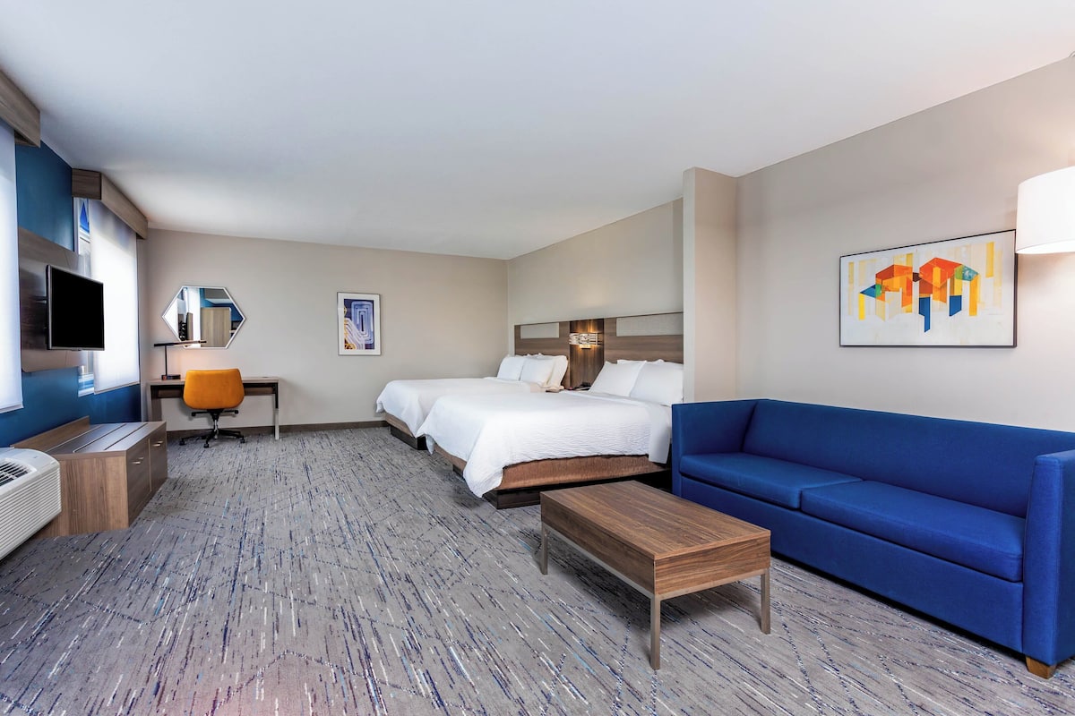 A hotel room with a bed, a blue sofa, a desk with a chair, a TV, and artwork on the walls.