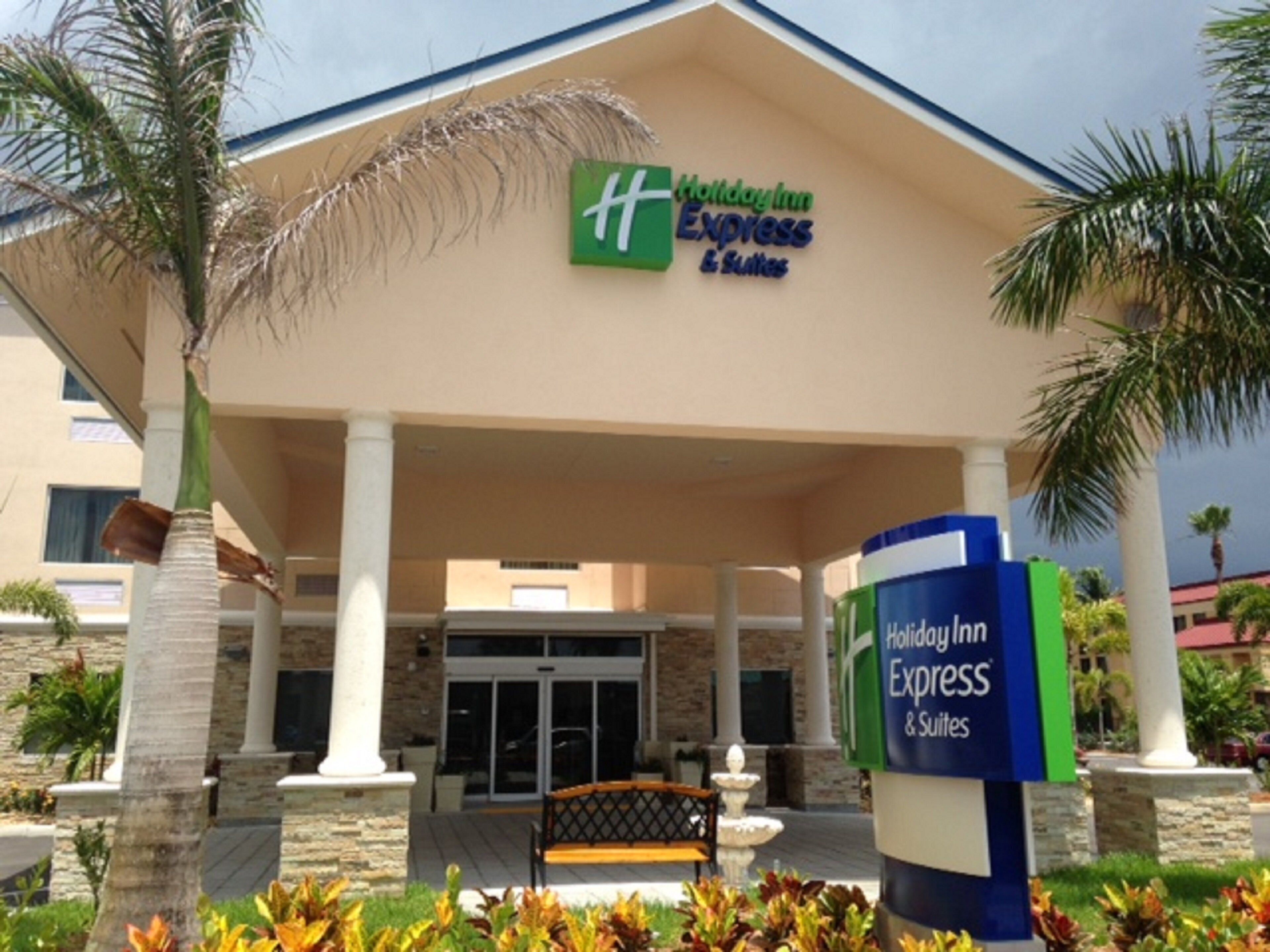 A Holiday Inn Express & Suites building with a covered entrance, palm trees, and a bench.
