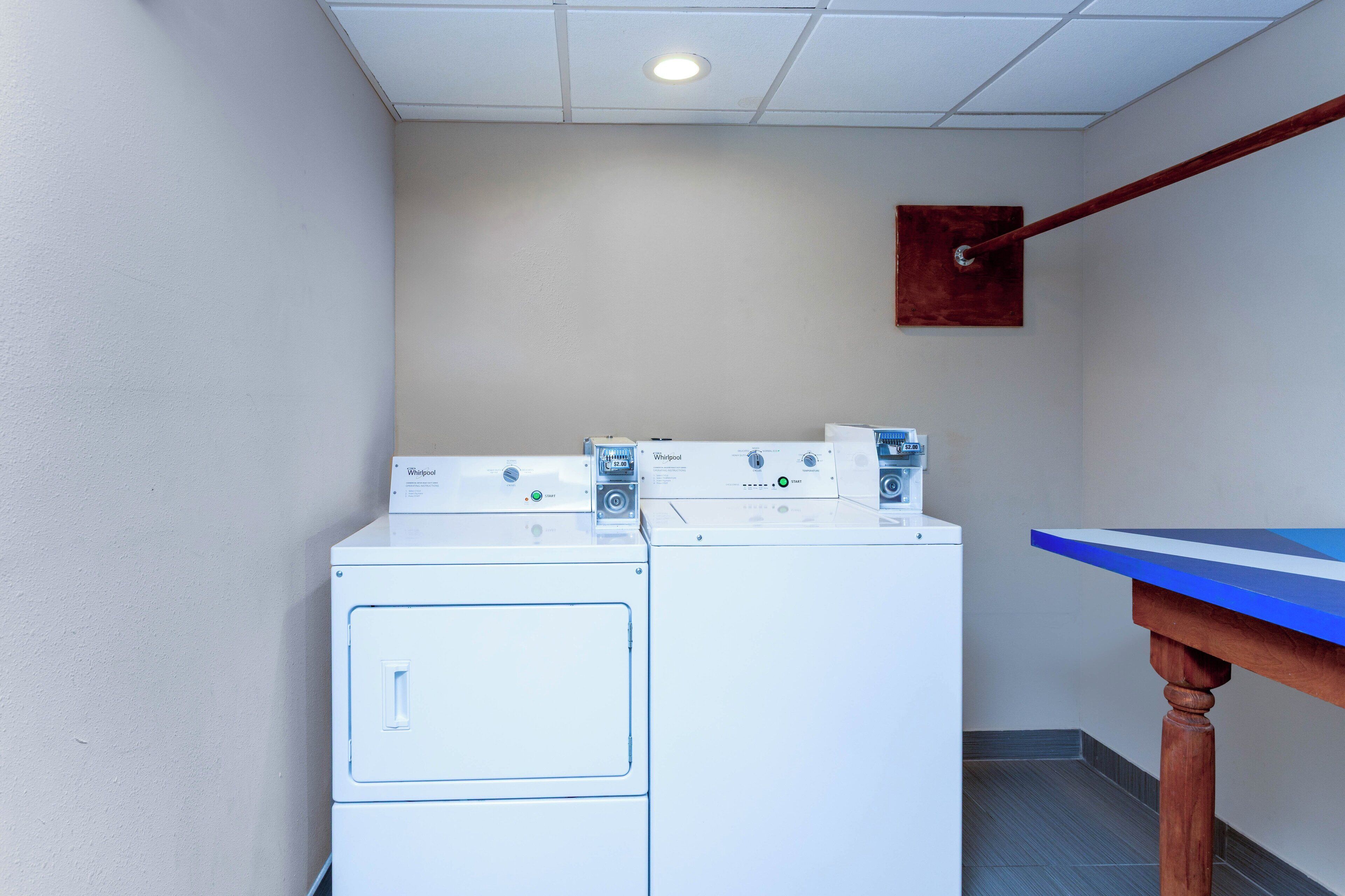 A laundry room with stacked washers and dryers, a blue table, and a wooden shelf.